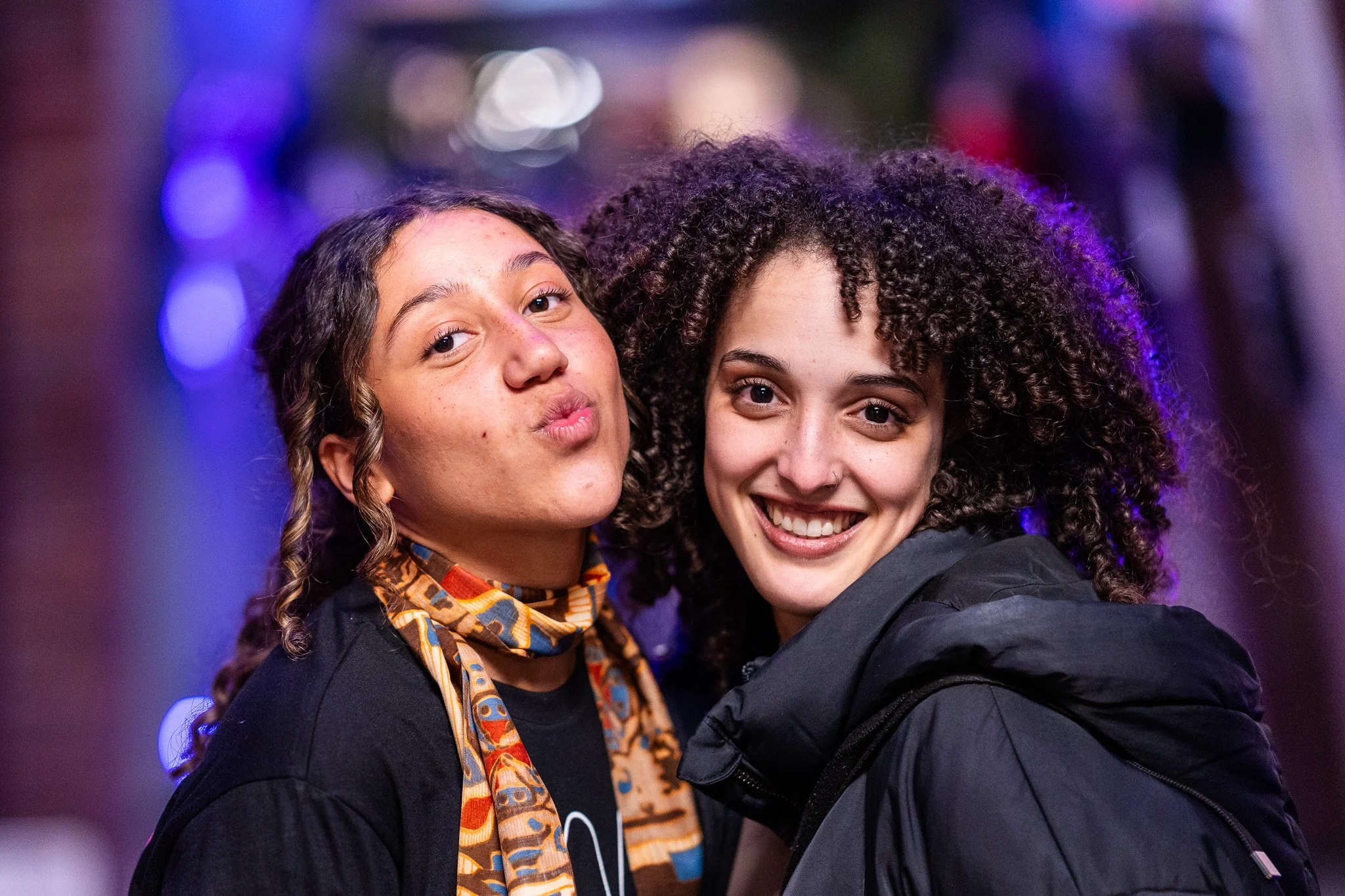 Two young women smiling and posing for a photo with blurred colorful lights in the background.