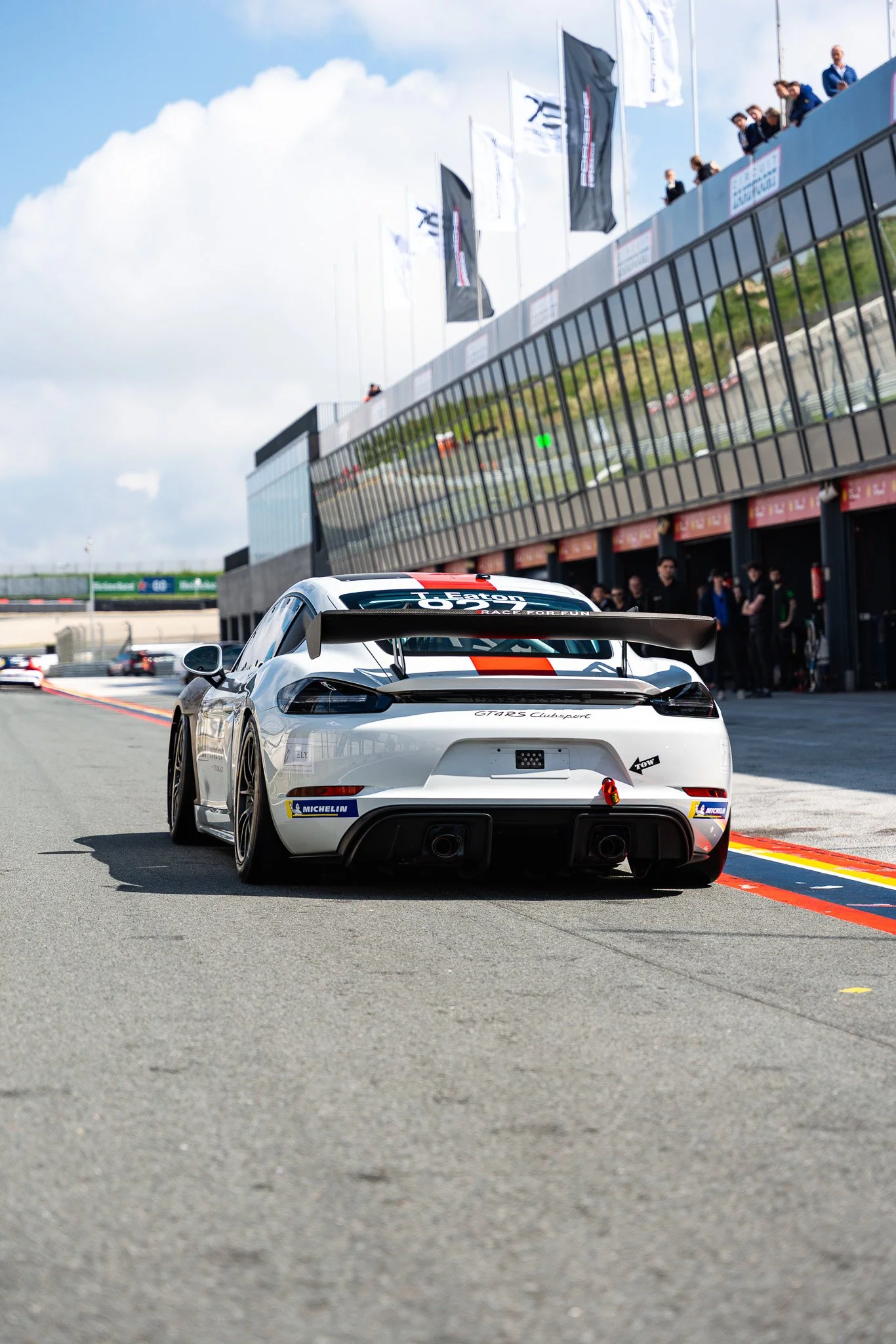 A white race car with a large rear wing on the race track during a racing event, with people observing from the upper level of the pit building and flags flying above.