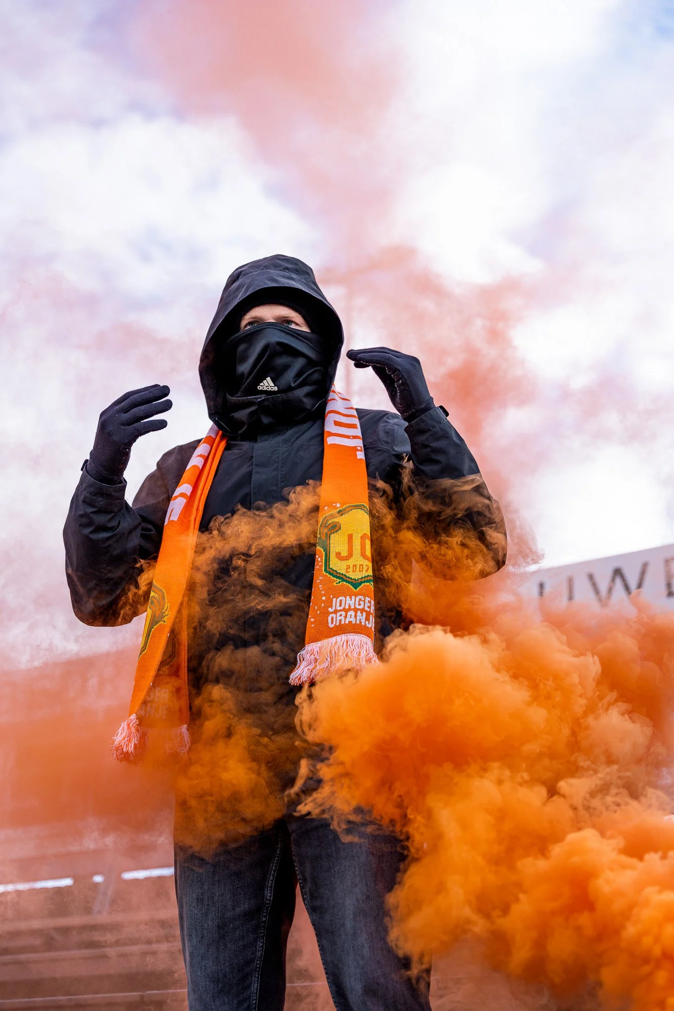 A person dressed in black winter clothing with a hood, face mask, and gloves, standing amidst orange smoke during a protest or demonstration, with a scarf that has orange, yellow, and green colors.