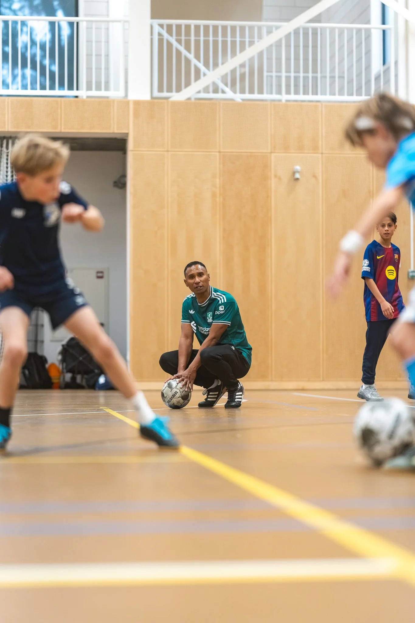 Kinderen spelen indoor volleyball met een coach die op de grond zit, in een sportschool met houten wanden en grote ramen.