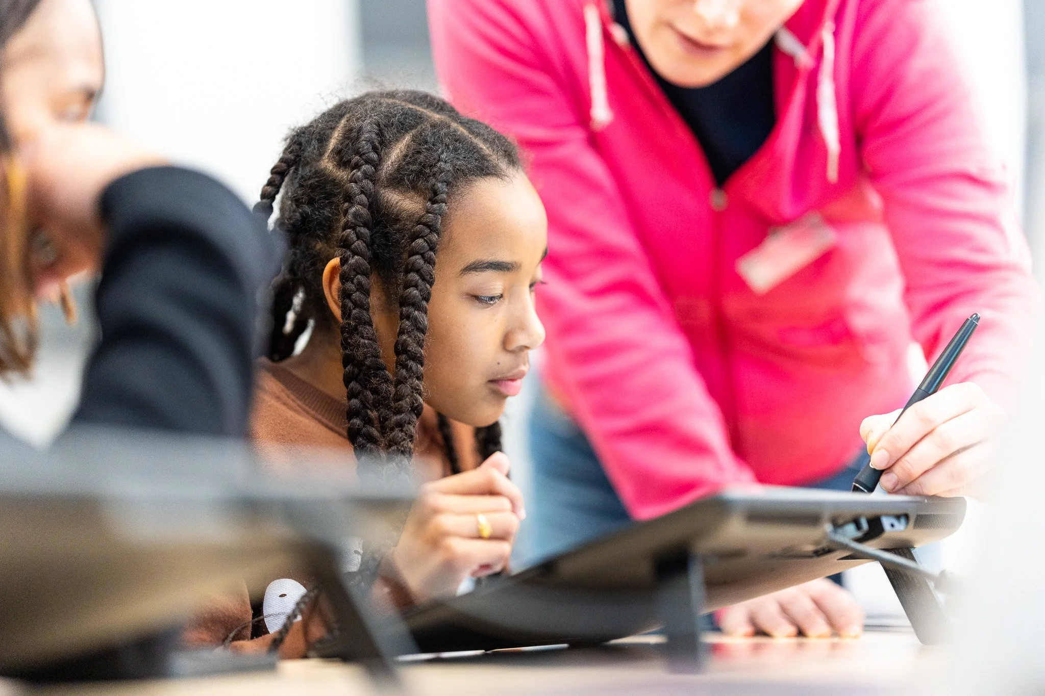 A young girl with braided hair looks at a computer tablet, while an adult woman in a pink hoodie points at the tablet with a pen, in a classroom or learning environment.