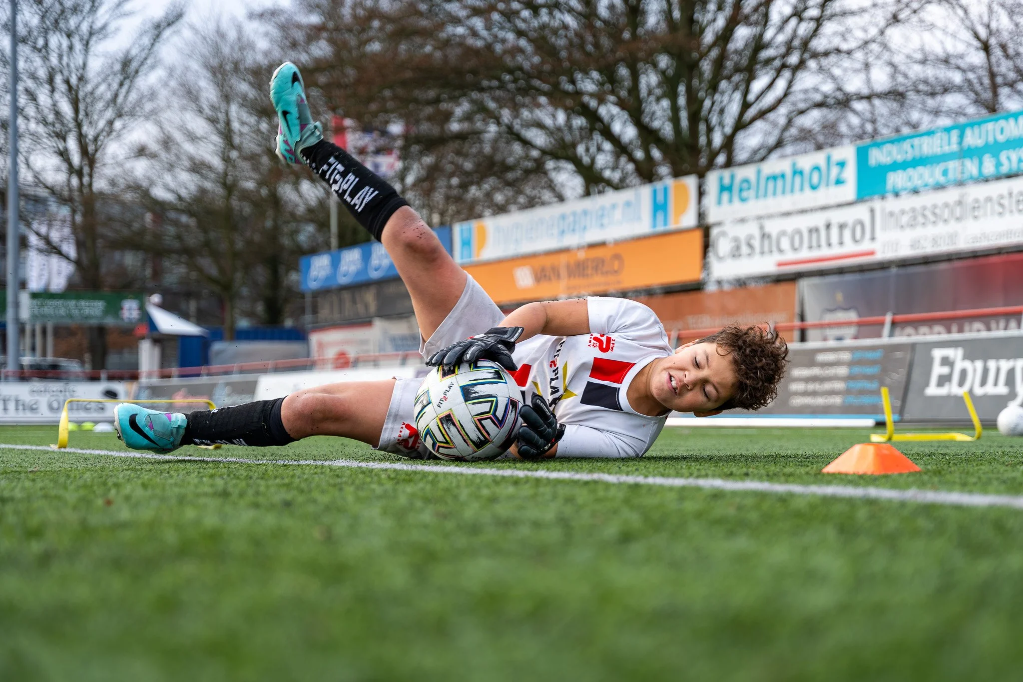 Een vrouwelijke keepster ligt op het veld en houdt een voetbal tegen haar lichaam, terwijl ze op haar zij ligt met één arm over de bal.