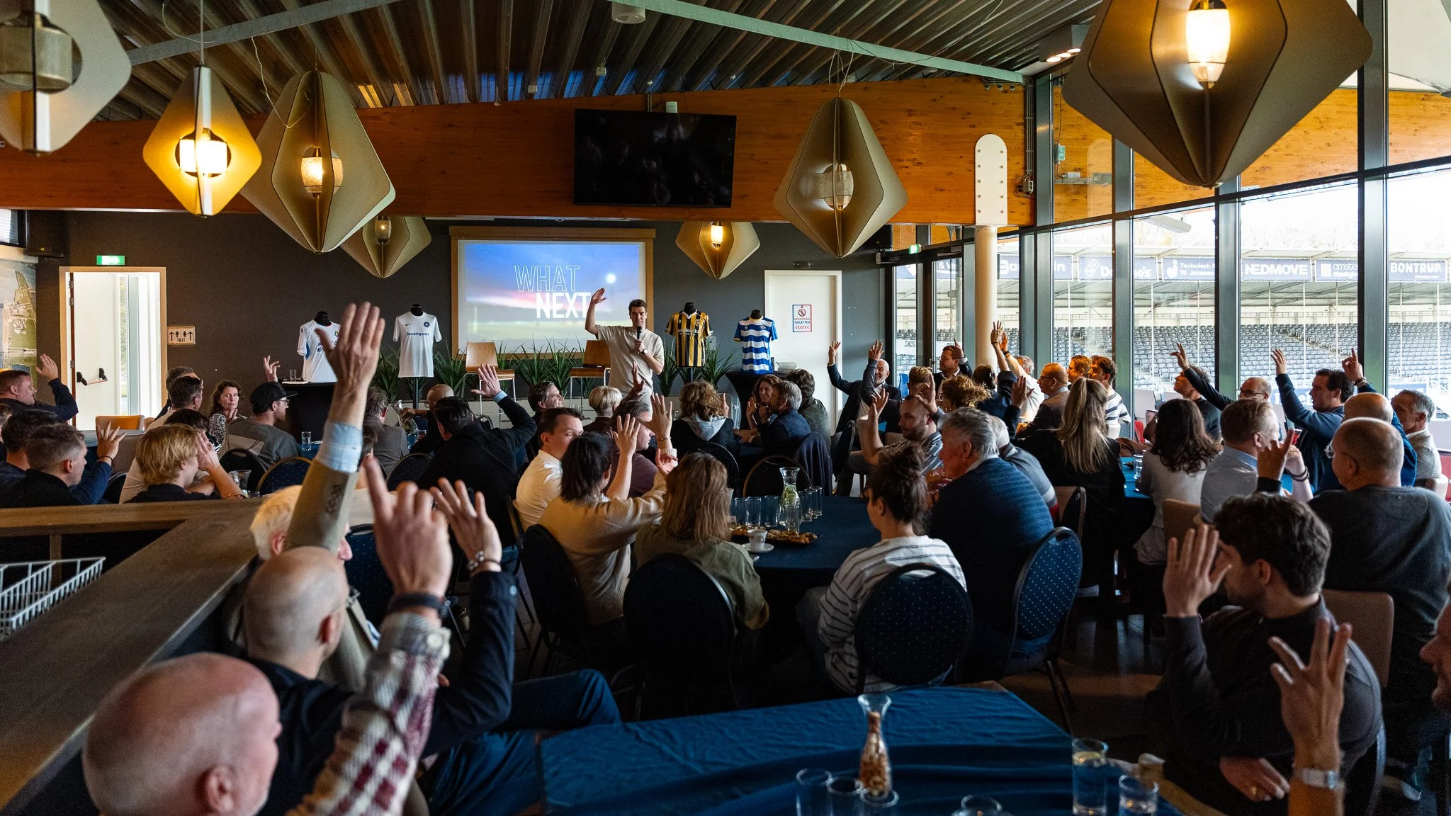 A man presenting at a conference with many attendees in a large, modern room, some raising hands.