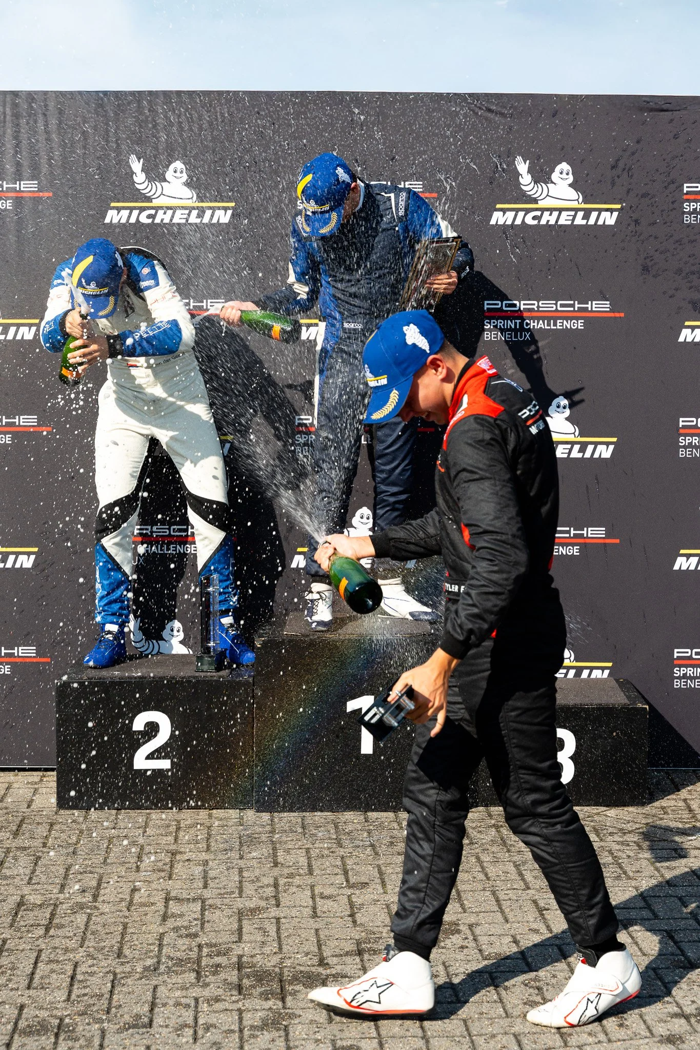 Race car drivers celebrating on a podium, spraying champagne in victory. One driver stands on the first-place position, while others are on second and third-place positions. The backdrop displays the Porsche Sprint Challenge Benelux logo and Michelin