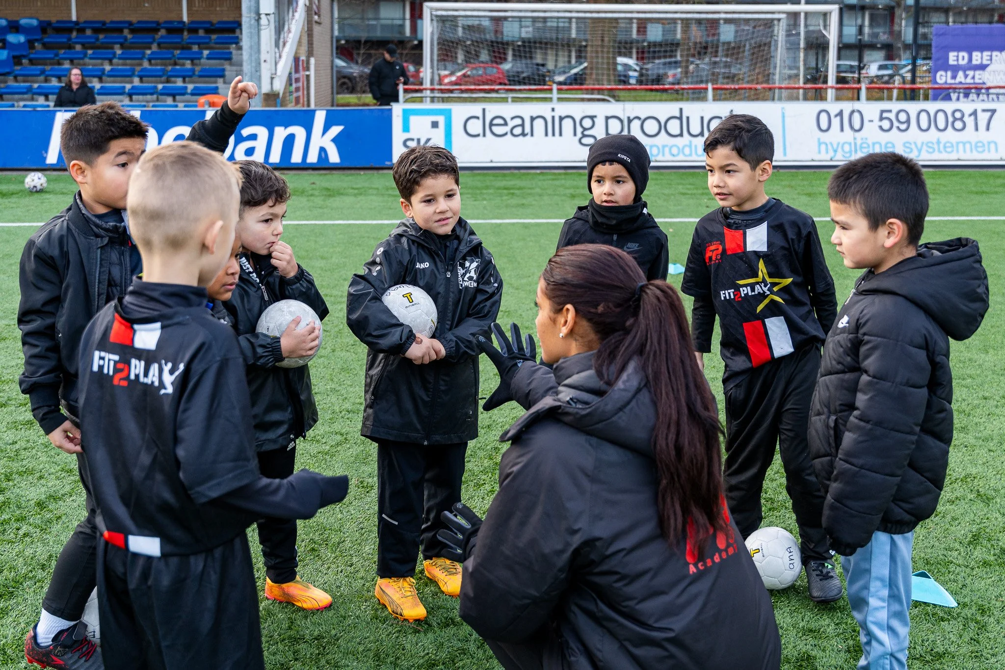 A women's coach giving instructions to a youth soccer team on a field. The team consists of young boys dressed in black training gear, holding soccer balls, and listening intently. The coach is kneeling and gesturing with her hands.