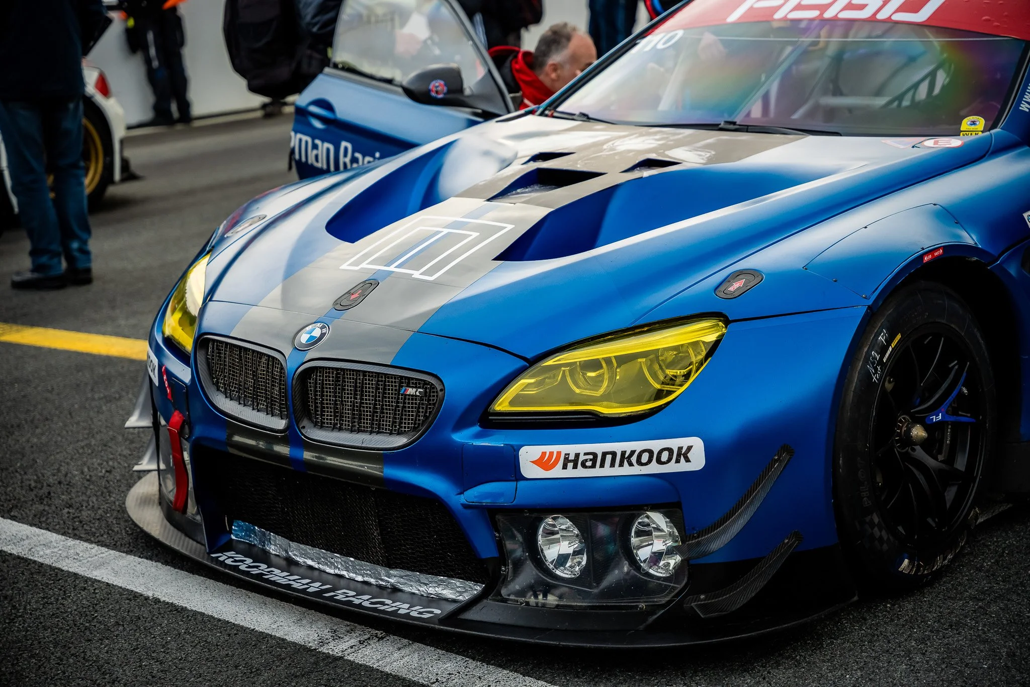 Blauwe raceauto met sticker 'Hankook' en geelgetinte koplampen, in pit lane met mensen en andere raceauto's op de achtergrond.