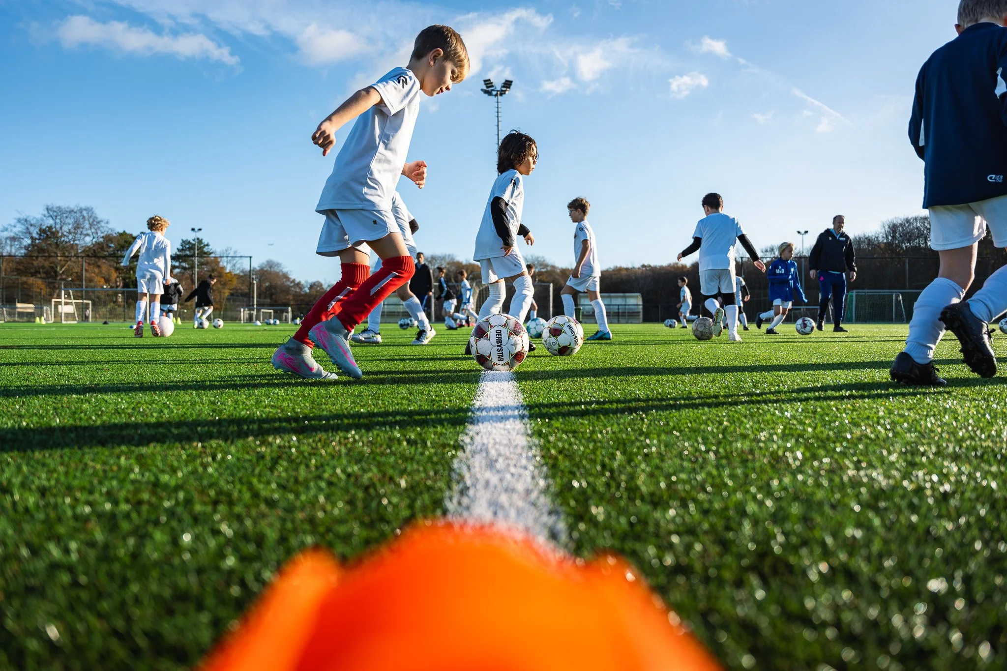 Kinderen trainen op een grasveld, met ballen en pionnen tijdens een jeugdvoetbaltraining op een zonnige dag.