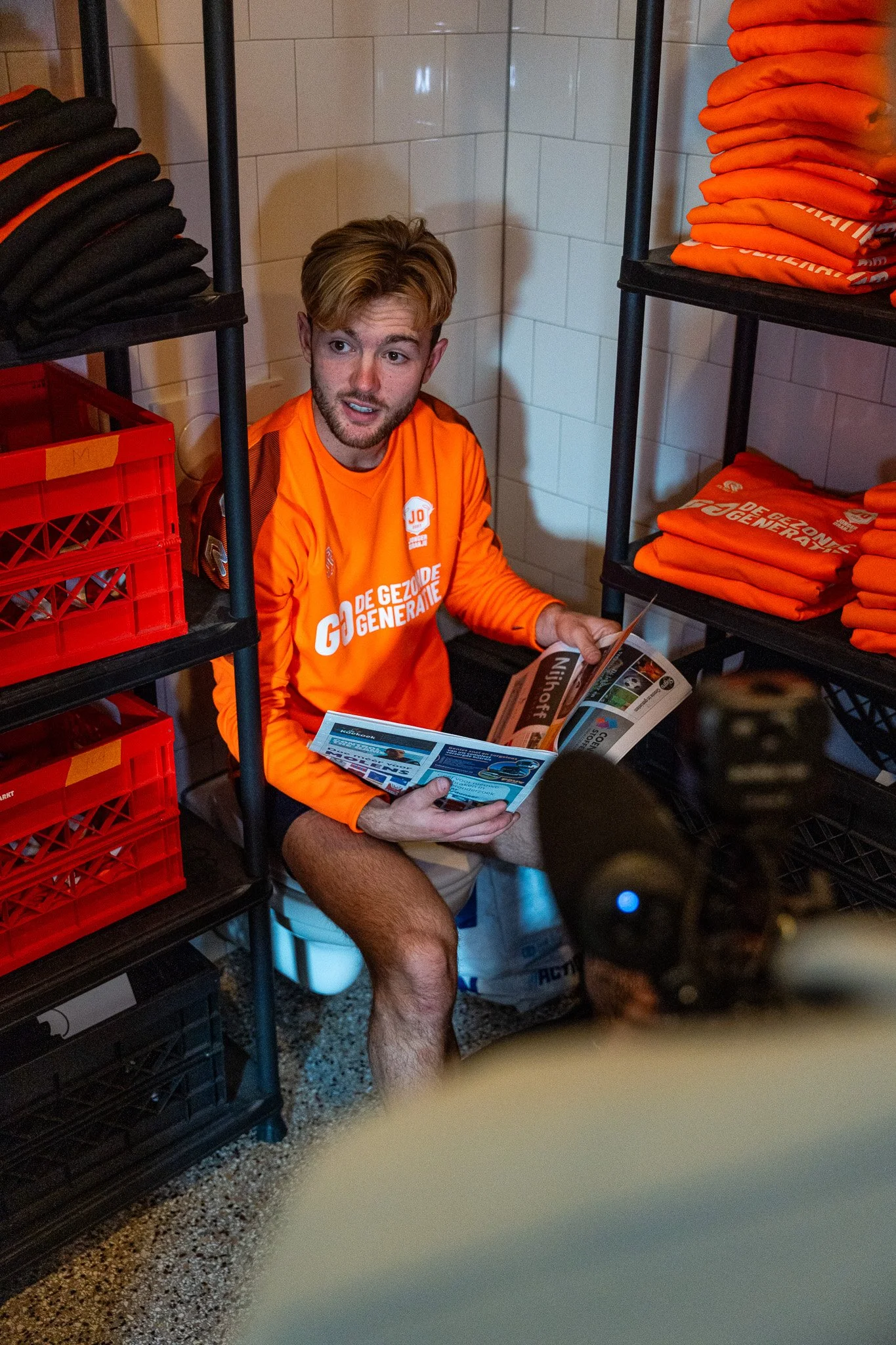 A young man with light skin, light brown hair, and facial hair, sitting on a toilet seat in a storage room, wearing an orange sports jersey with white text and logos, holding a magazine, surrounded by shelves holding orange and black clothing and red