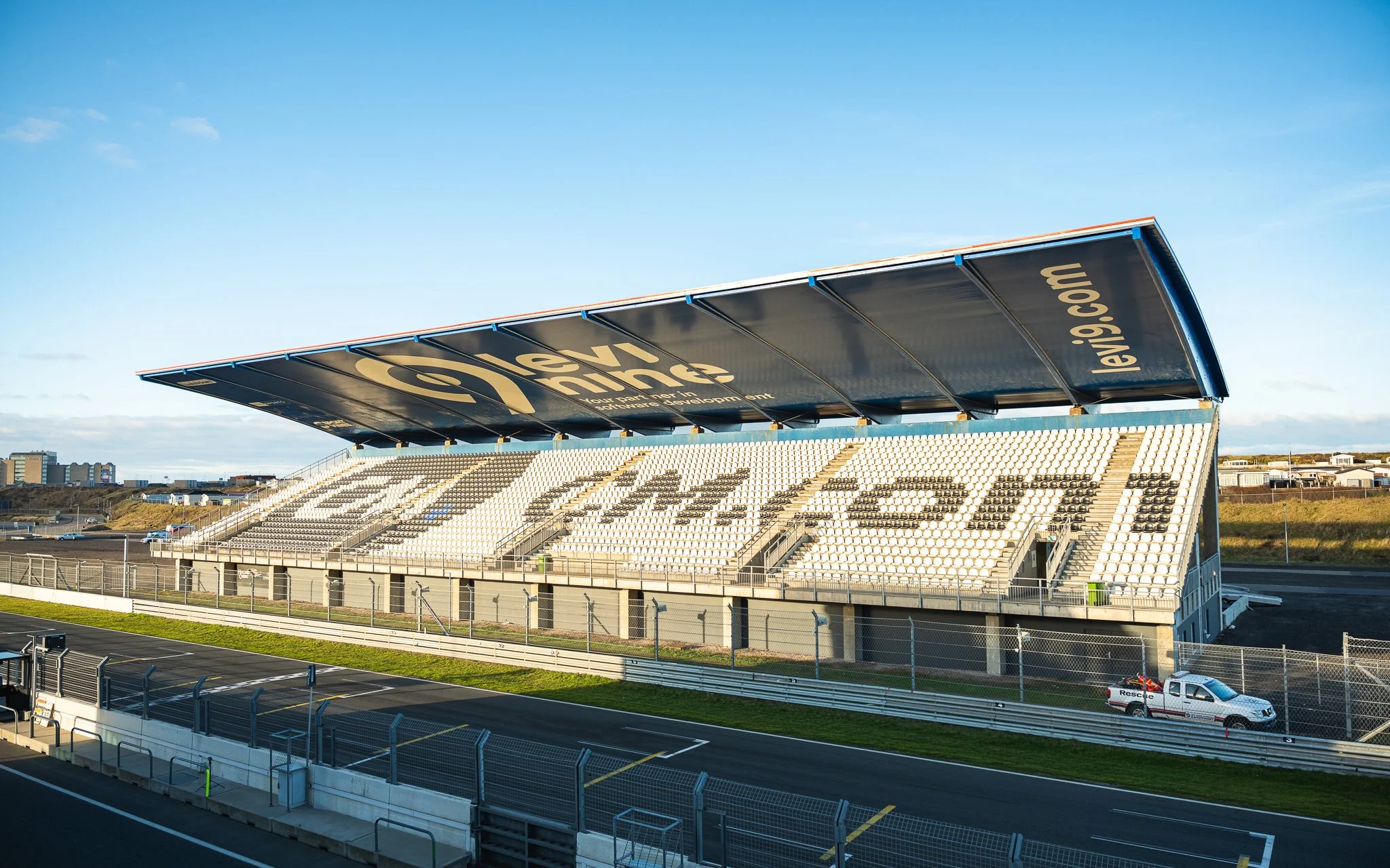 Empty race track grandstand with large blue canopy displaying 'leap9.com' and 'race' on seats, with rescue vehicle parked nearby under a blue sky.