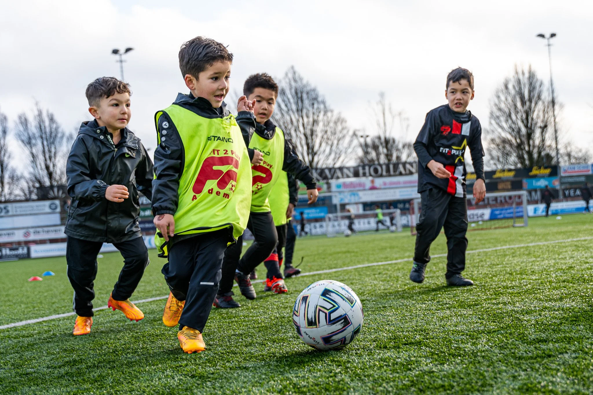 Vijf jonge jongens die op een voetbalveld oefenen, met enkele in het zwart en twee in gele trainingspakken.