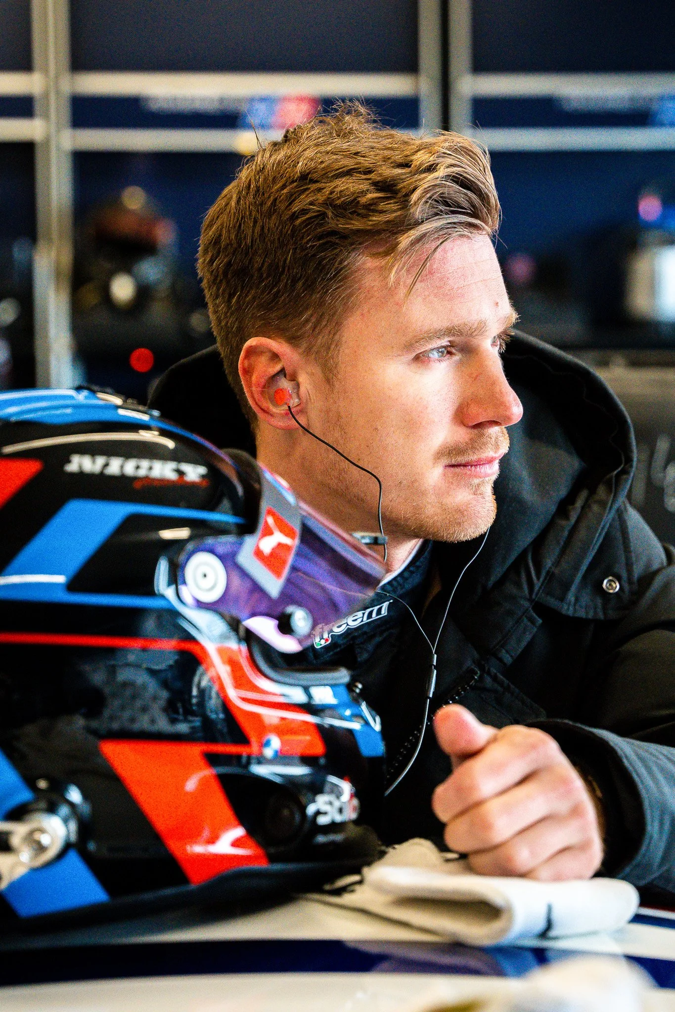 A race car driver with a blue, red, and black helmet resting on a table, wearing earphones and a black jacket, looking to the right.