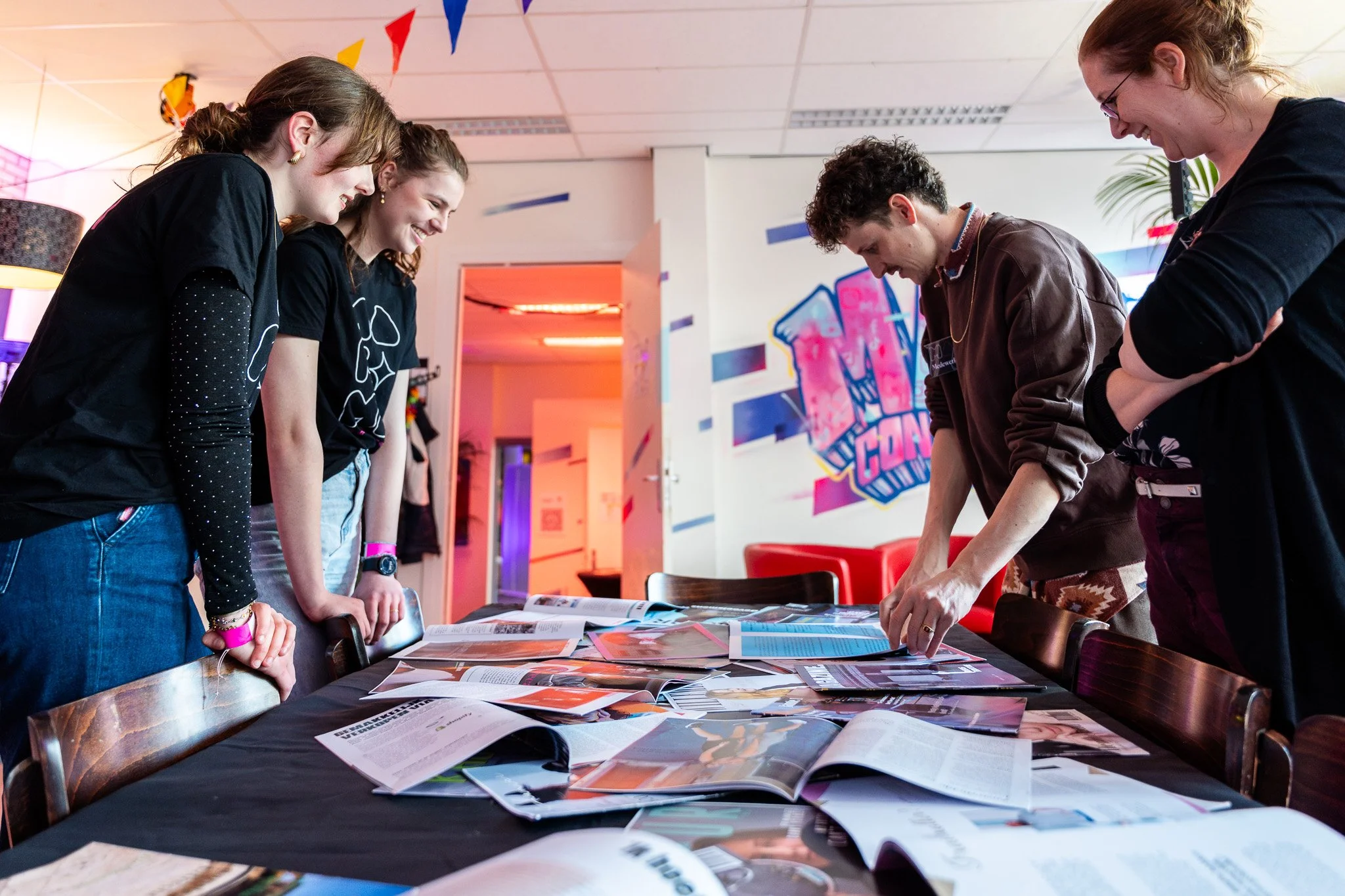 Four young adults are gathered around a table covered with magazines and papers, looking at materials and engaging in discussion in a brightly lit, colorful room with graffiti-style wall art.