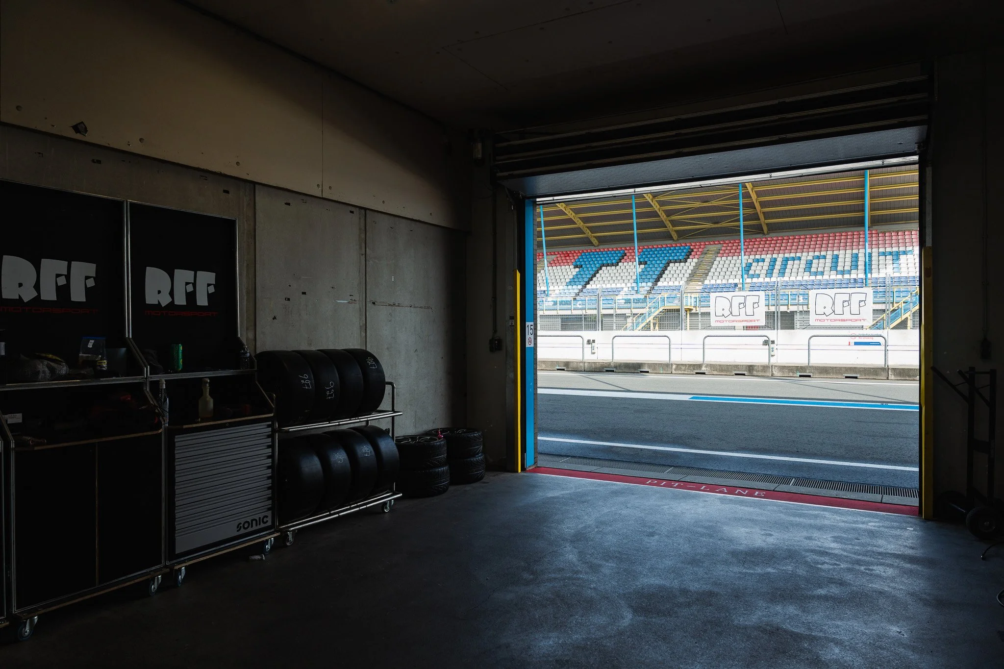 View from inside a race garage looking out through an open door to a race track with stadium seating and banners that say 'RFF Motorsport'.