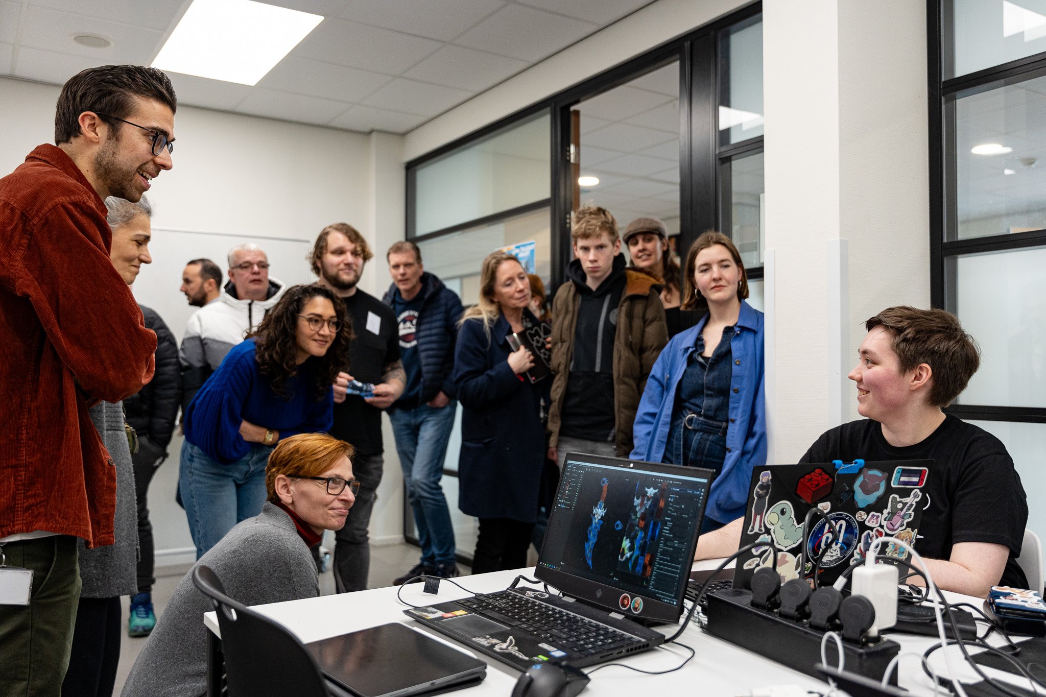 A group of people in a computer lab gathered around a young man sitting at a desk with a gaming computer and monitors, listening to him speak.
