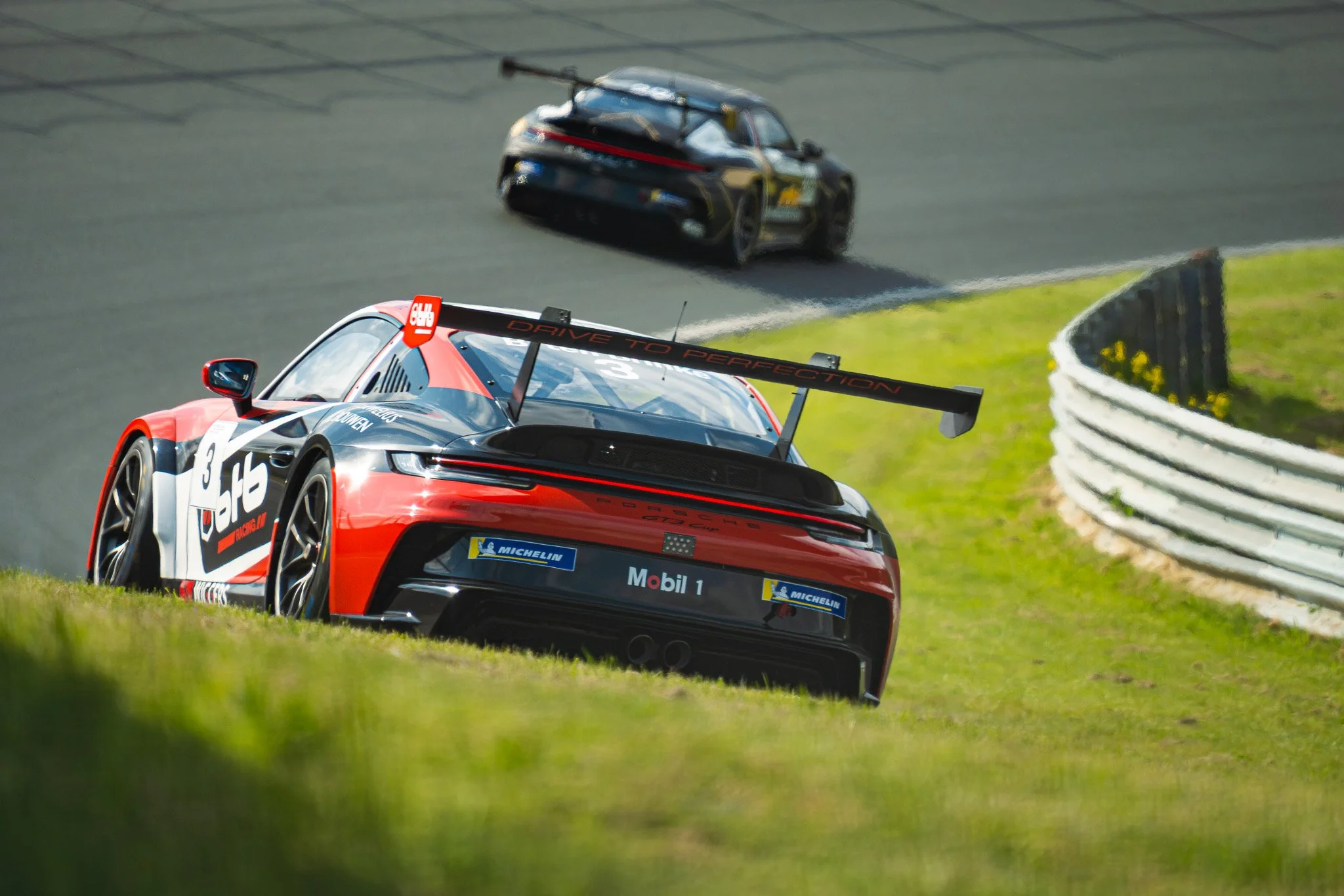 Two race cars on a track, one in the foreground with red, black, and white livery and sponsorship stickers, including Porsche branding, and another in the background with a black and blue design, navigating a bend on the circuit.