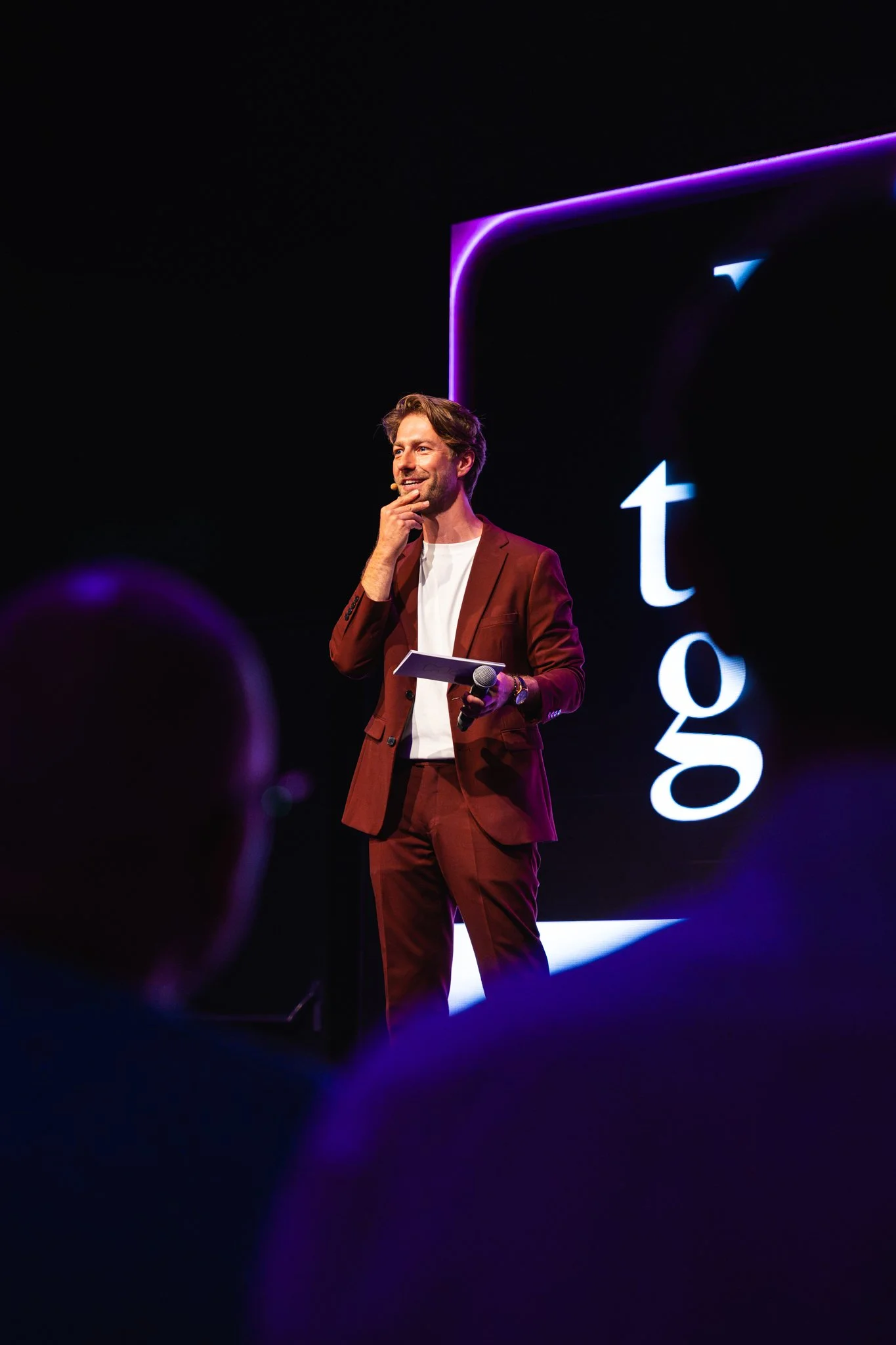 Man in een bruin pak met witte T-shirt staat op een podium met een microfoon in de hand, voor een groot scherm met het Google logo en neonachtige lichtaccenten.