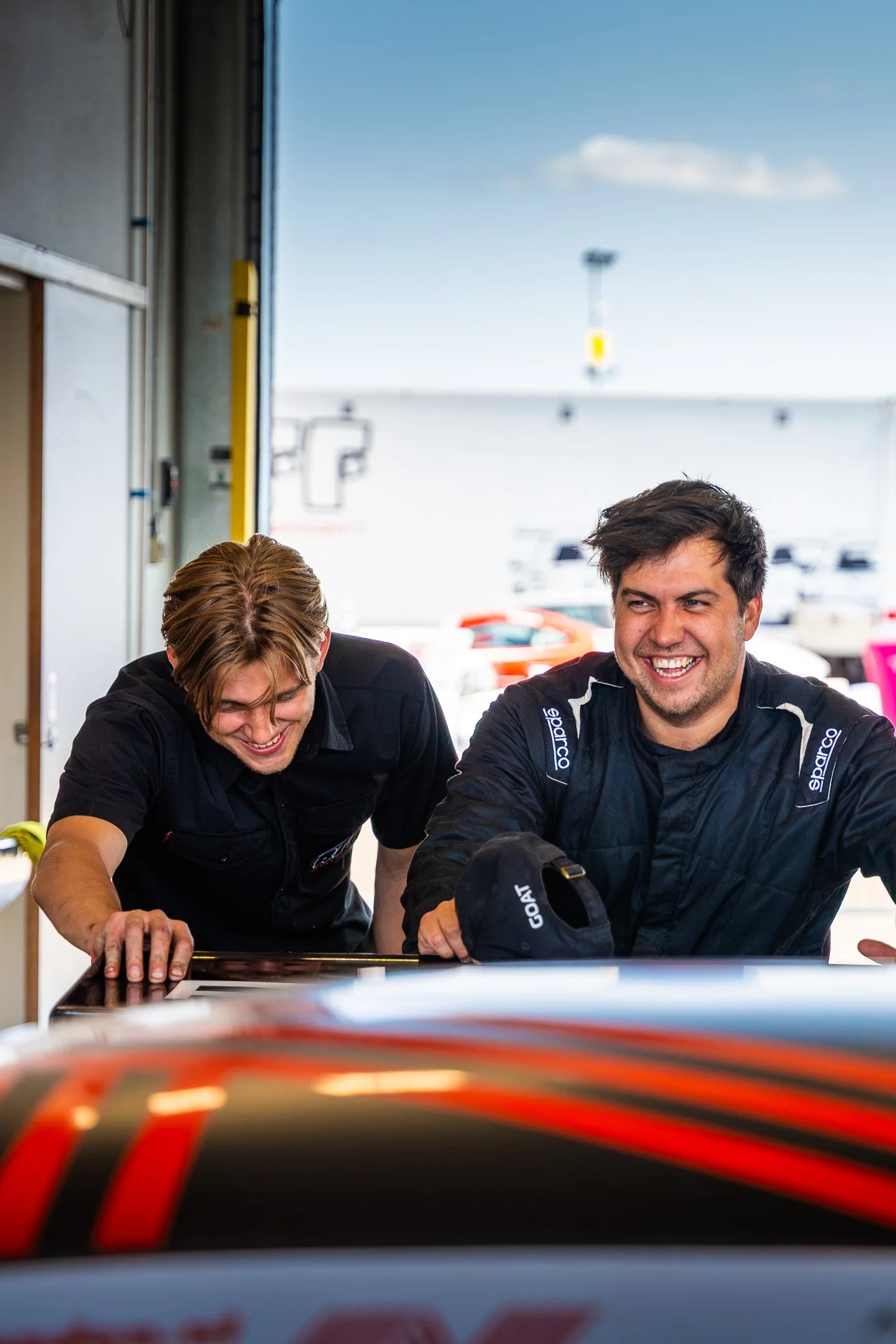 Twee mannen lachen en kijken naar iets op een tafel in een garage met raceauto's op de achtergrond.