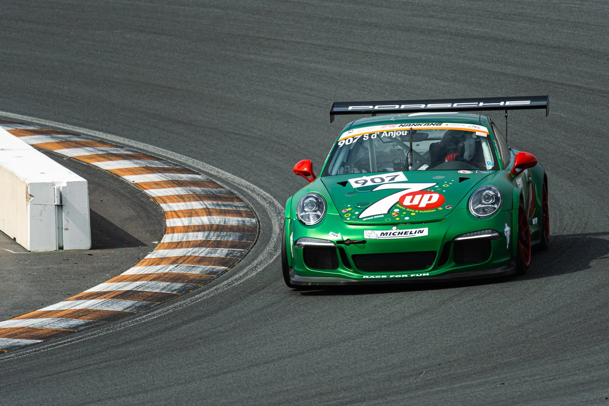 Groene Porsche raceauto op een circuit, inclusief racestickers en een grote achtervleugel, neemend een bocht met gele en oranje markeringen.