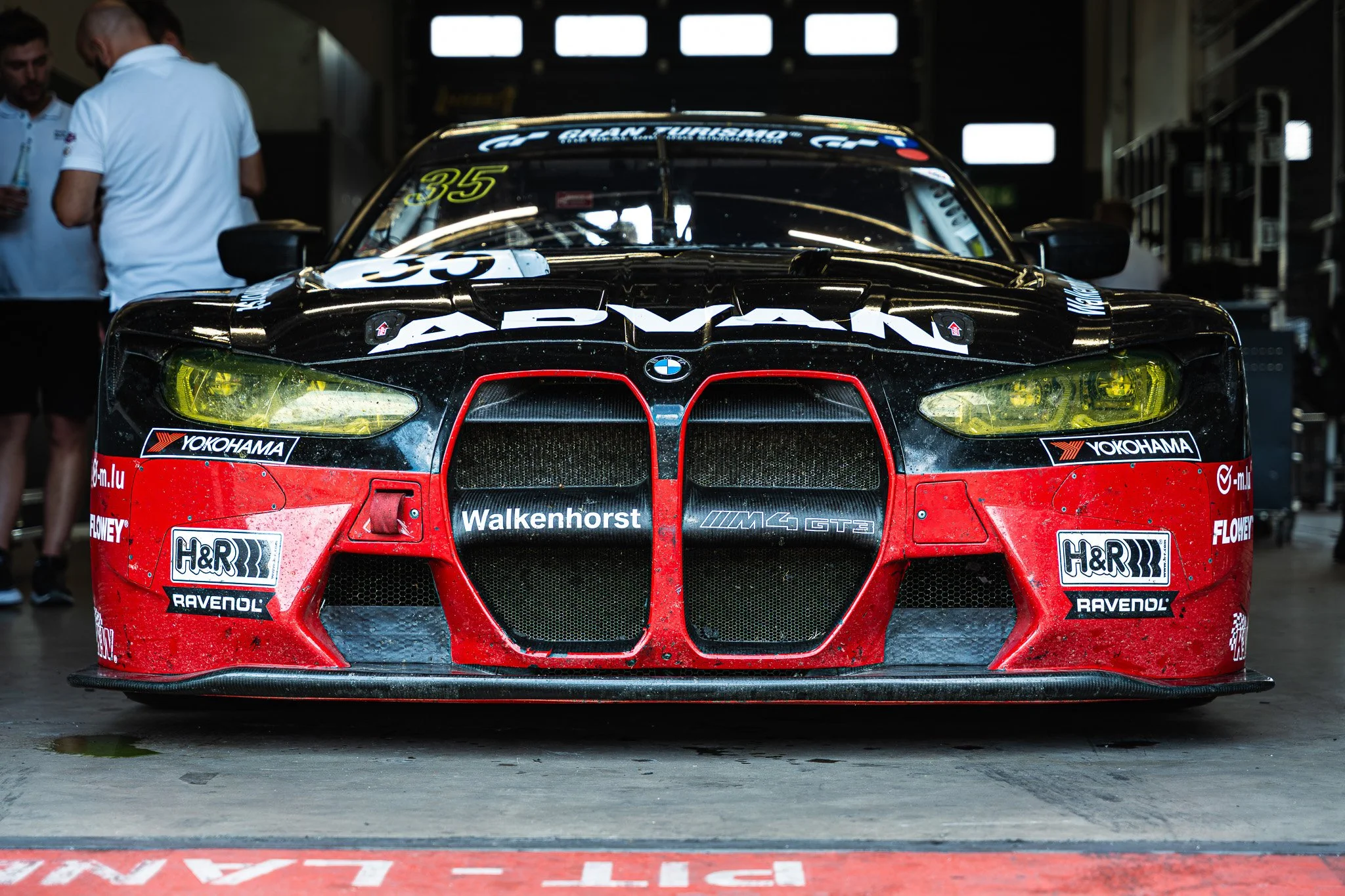 Front view of a black and red BMW race car with yellow headlights, sponsored by Yokohama, H&R, and Ravenol, parked in a garage with team members in the background.