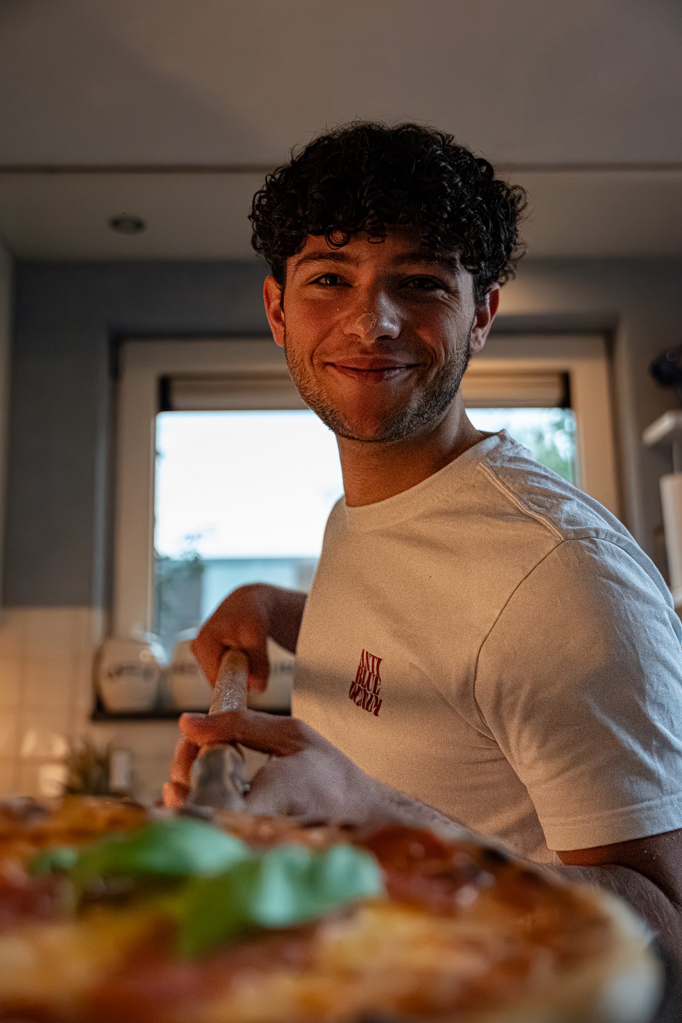 A smiling man with curly hair in a kitchen, holding a pizza with vegetables. The background shows a window and kitchen shelves.