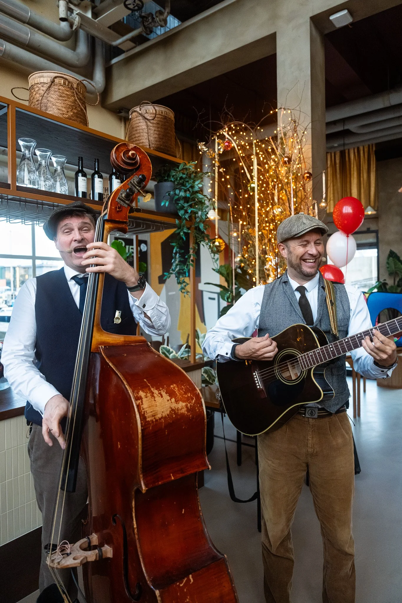 Two men happily performing music in a cozy, decorated indoor space. One is playing a double bass, and the other is playing an acoustic guitar. The background features warm lighting, plants, and balloons, indicating a festive or celebratory event.