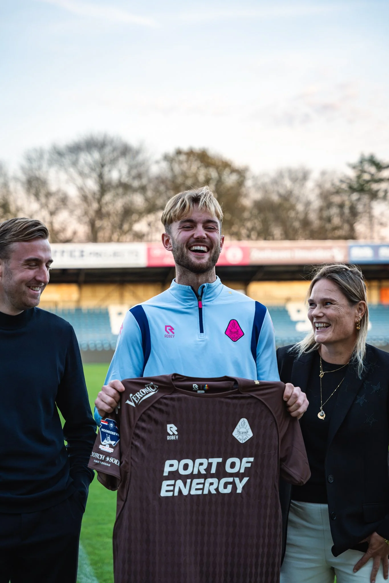 Vijf mensen, drie vrouwen en twee mannen, staan op een voetbalveld en lachen. Een man draagt een voetbalshirt en houdt een teamtrui met tekst "Port of Energy". De sfeer is vrolijk en de achtergrond toont bomen en een stadion.