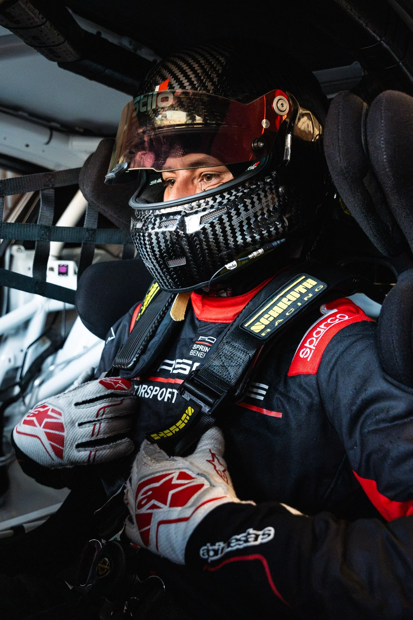 Close-up van een raceautocoureur in een raceauto, met een helm en handschoenen, gekleed in een jumpsuit met sponsorlogo's.