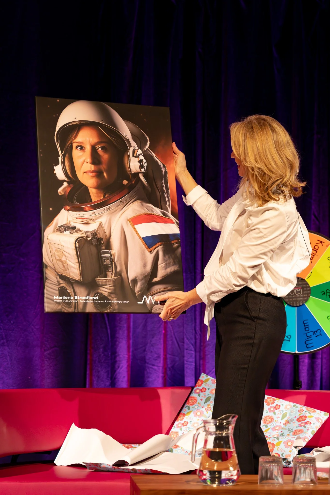 Vrouw met blonde haar hangt een grote poster van een astronaut in een ruimtepak op aan een muur met een paarse achtergrond.
