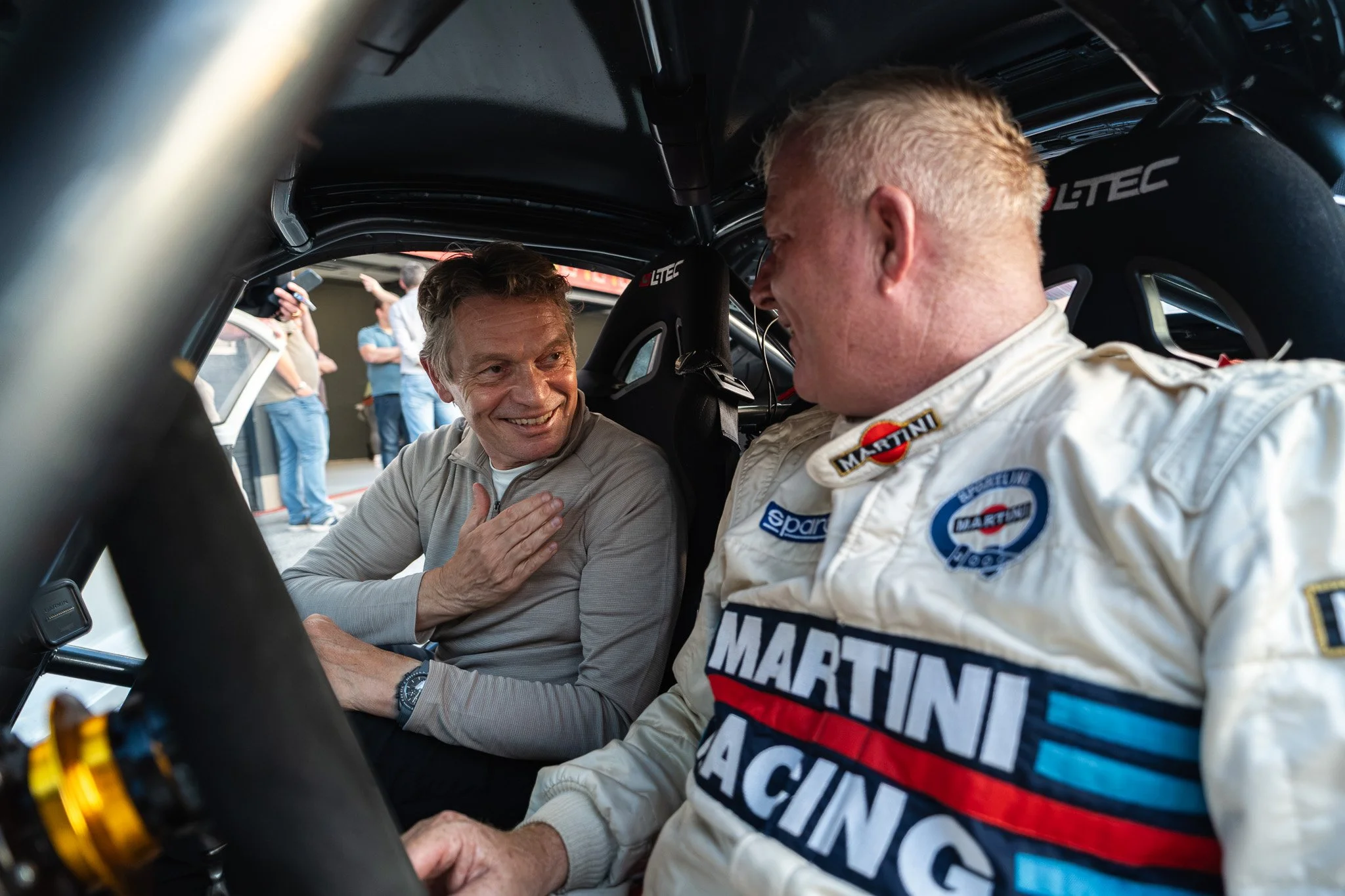 Twee mannen in een racewagen, een glimlachende man in een beige trui en een man in een witte raceoutfit met merken als Martin Engineering en Sparco, praten en schudden handen binnen de auto. Mensen staan buiten en kijken toe op de achtergrond.