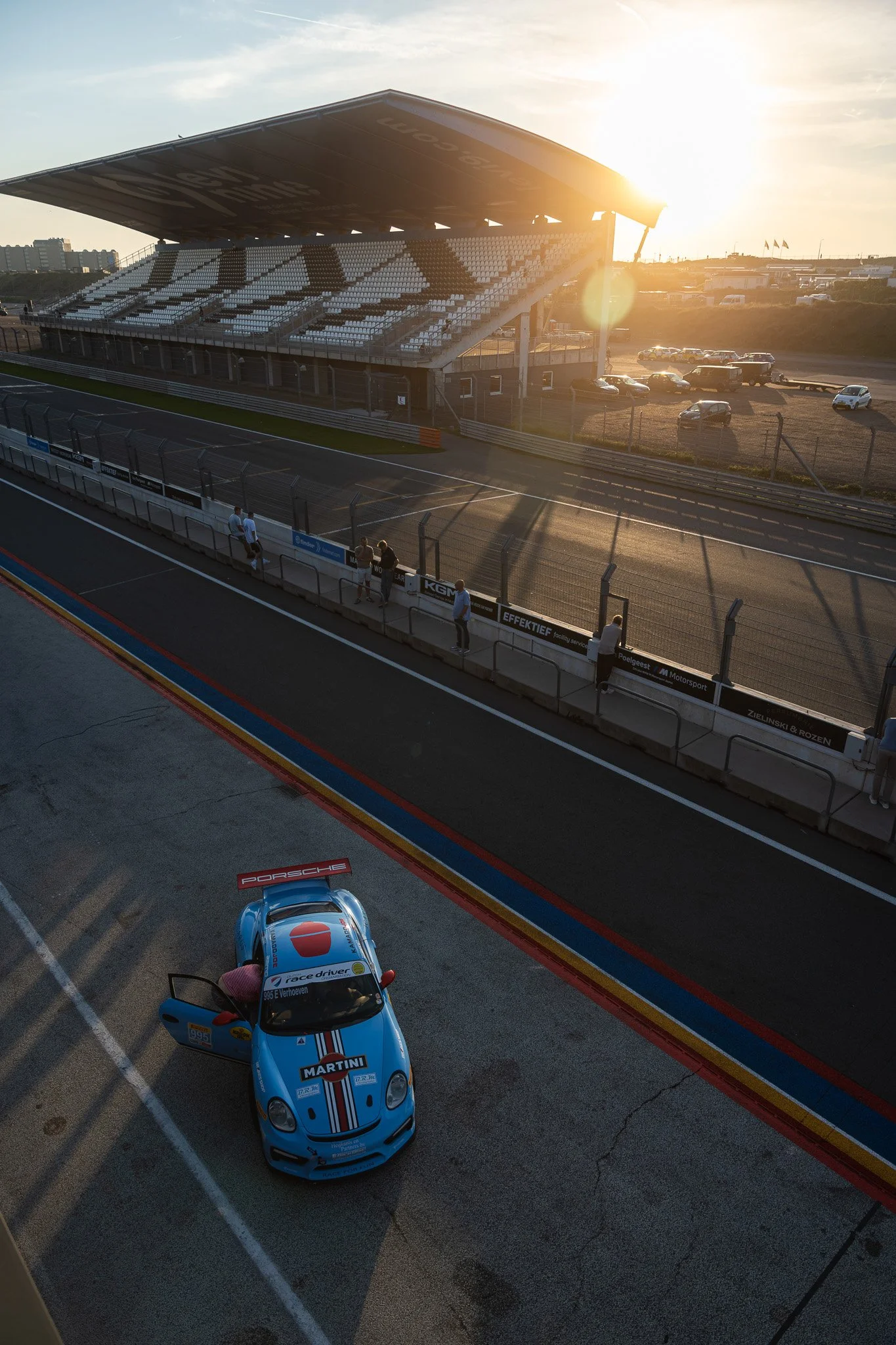 Raceauto met Martini-livery op de pitbox bij een racecircuit in de namiddag, met enkele mensen en een parkeerplaats op de achtergrond.