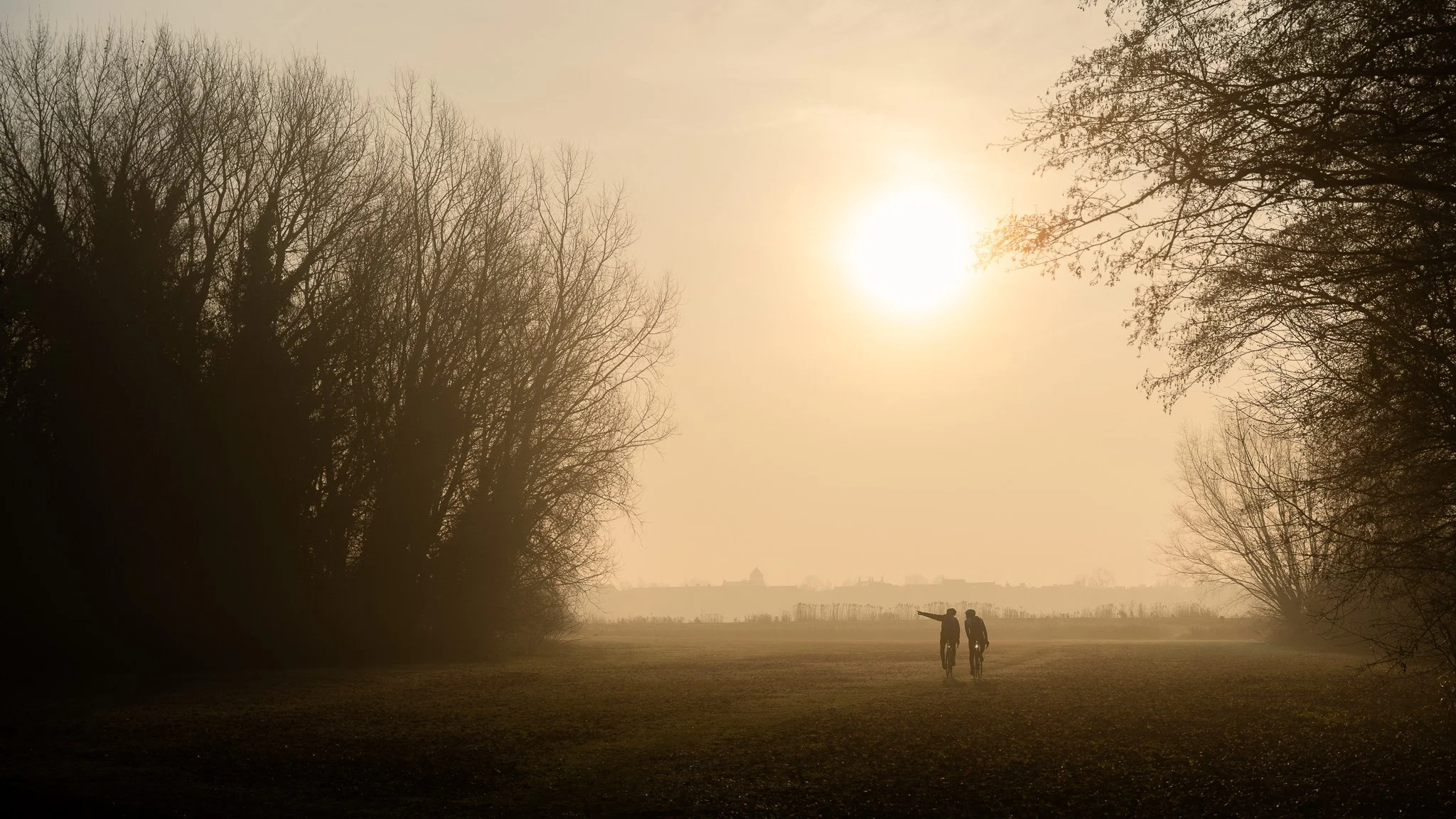Two people, possibly children, walking on a grassy field during sunrise or sunset, with trees on either side and a hazy distant landscape.