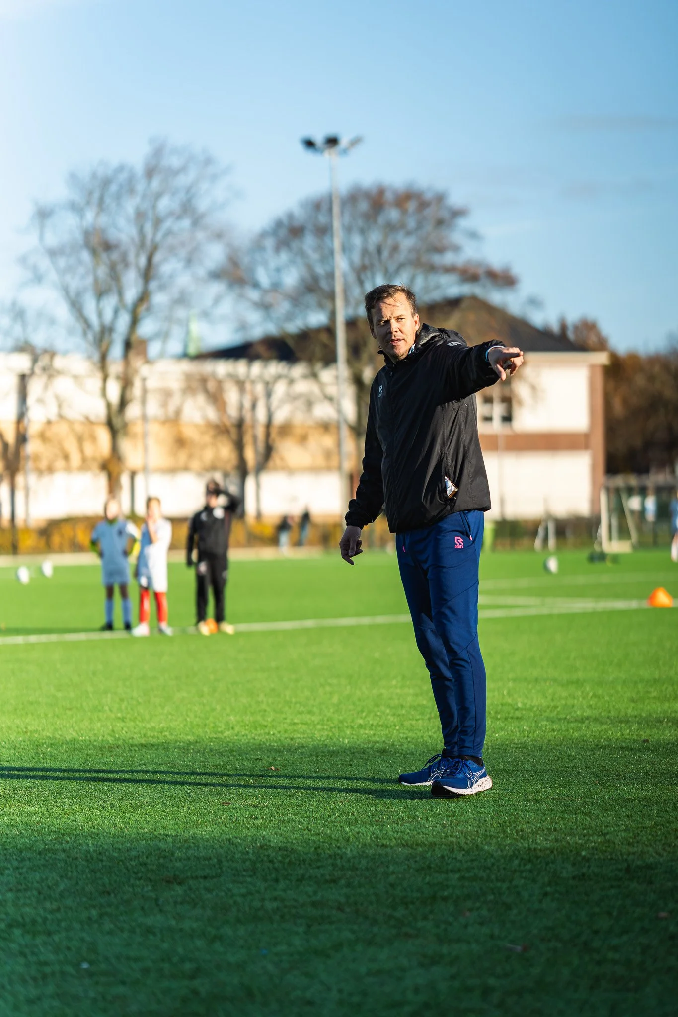 Een man in sportkleding waarschuwt op een grasveld met voetbaldoelen, omringd door kinderen tijdens een buitensportactiviteit.