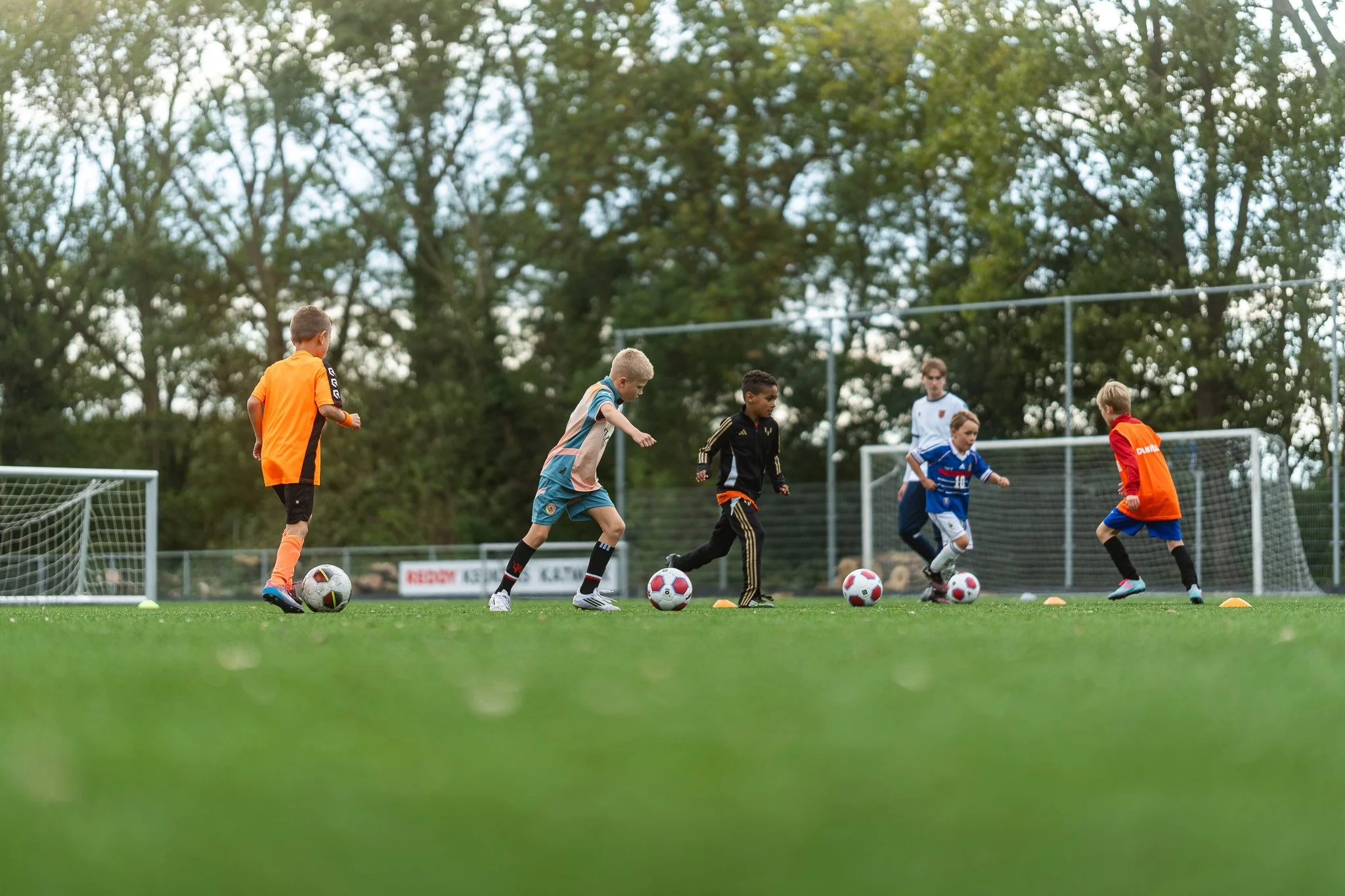 Jonge jongens die voetbal oefenen op een grasveld met een hek en bomen op de achtergrond.