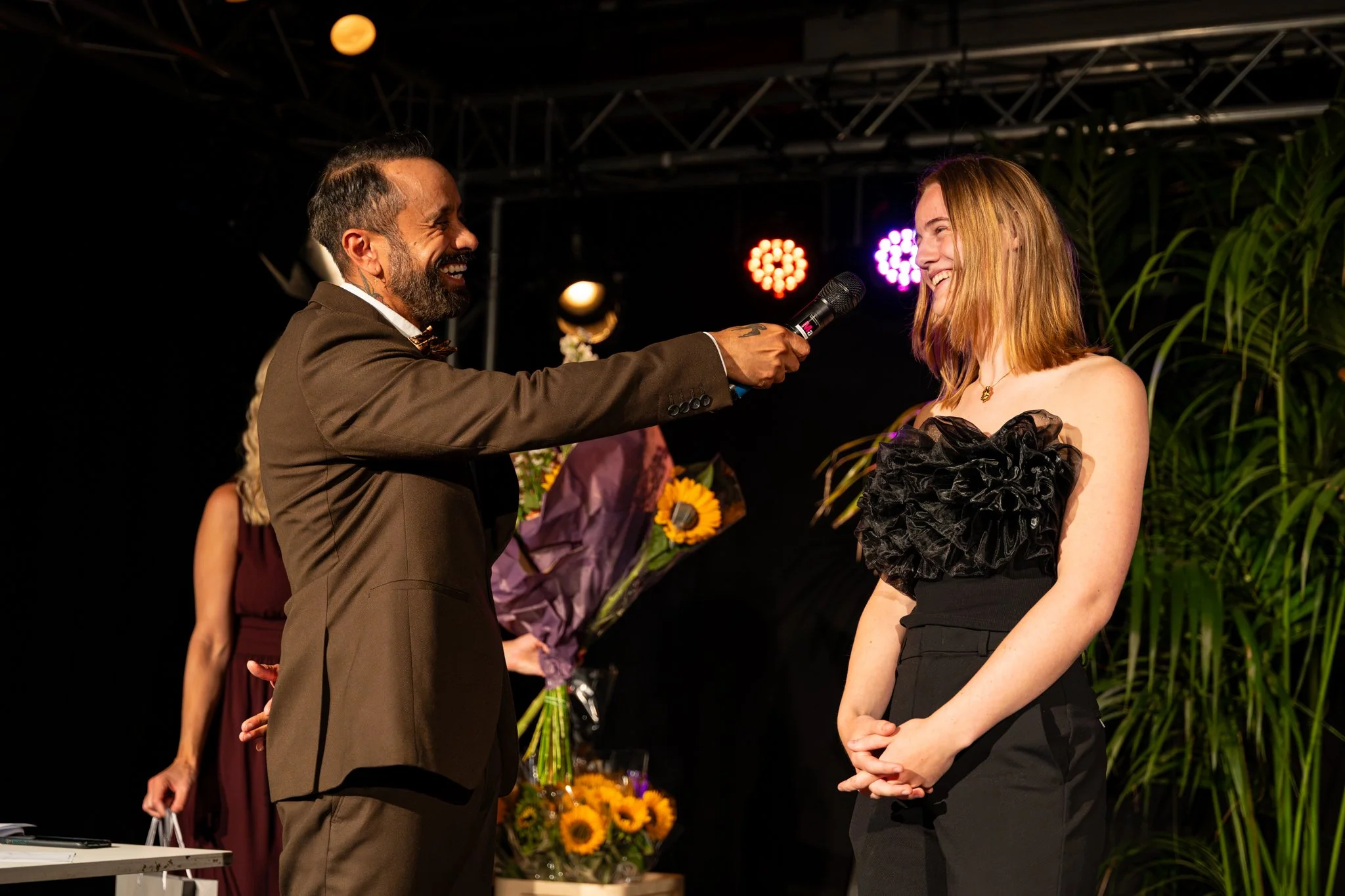Man met een beard en donker pak houdt een microfoon richting een glimlachende jonge vrouw met rood haar en een zwart jurk op een feestelijk evenement. Op de achtergrond zijn bloemen en gekleurde lichten zichtbaar.