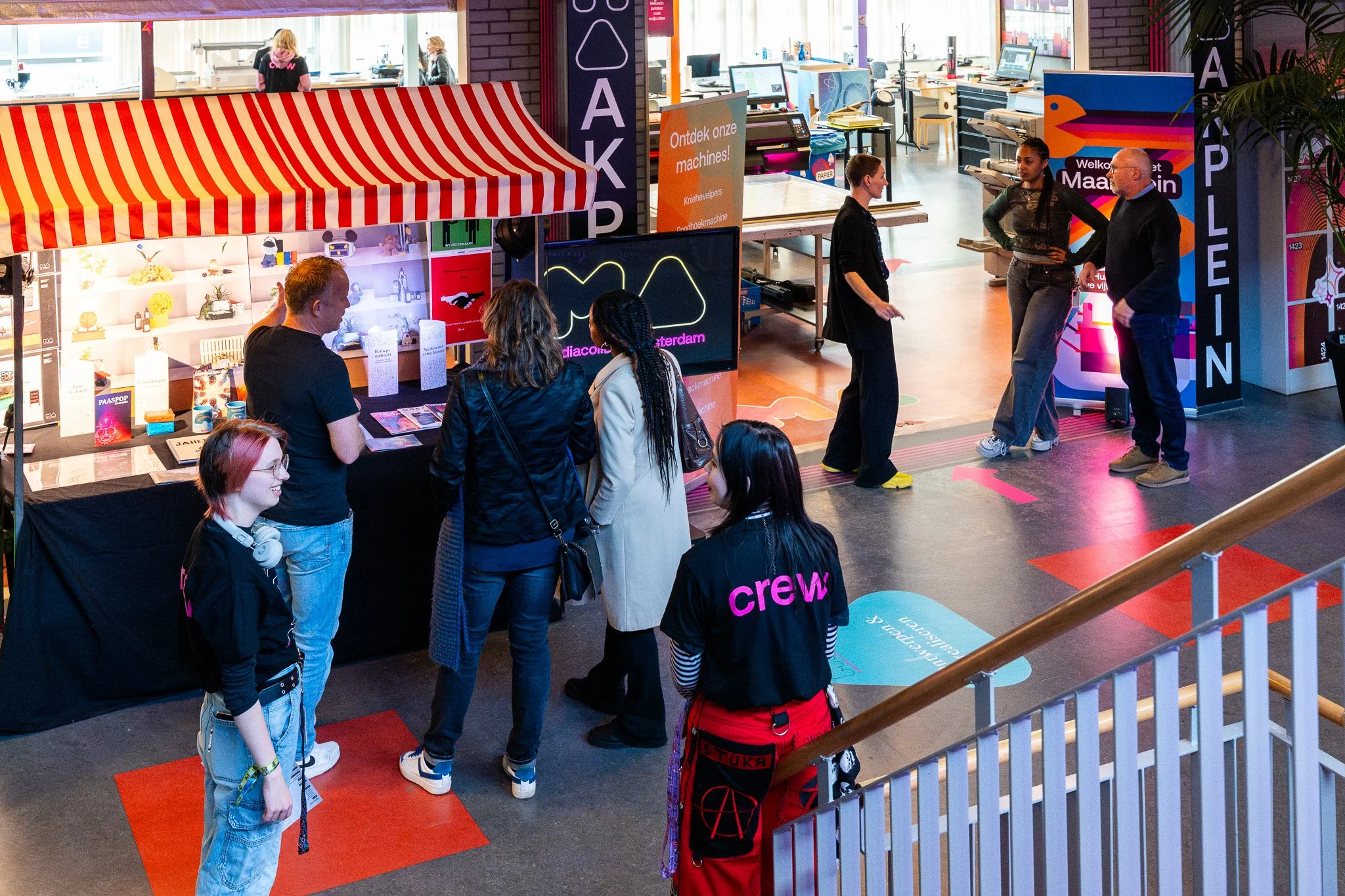 People gathered around a booth at an indoor event, with some talking to staff behind the booth and others waiting in line. The booth has a striped red and white canopy and displays products, with screens and banners in the background.