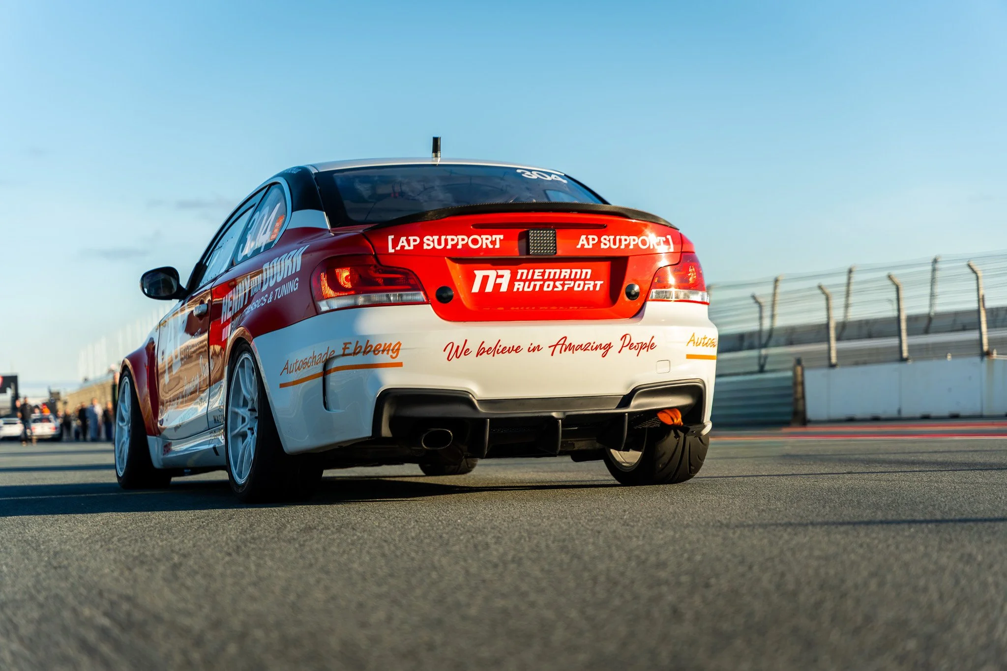 Wit en rood raceauto op een circuit met een blauwe lucht en een omheining op de achtergrond.