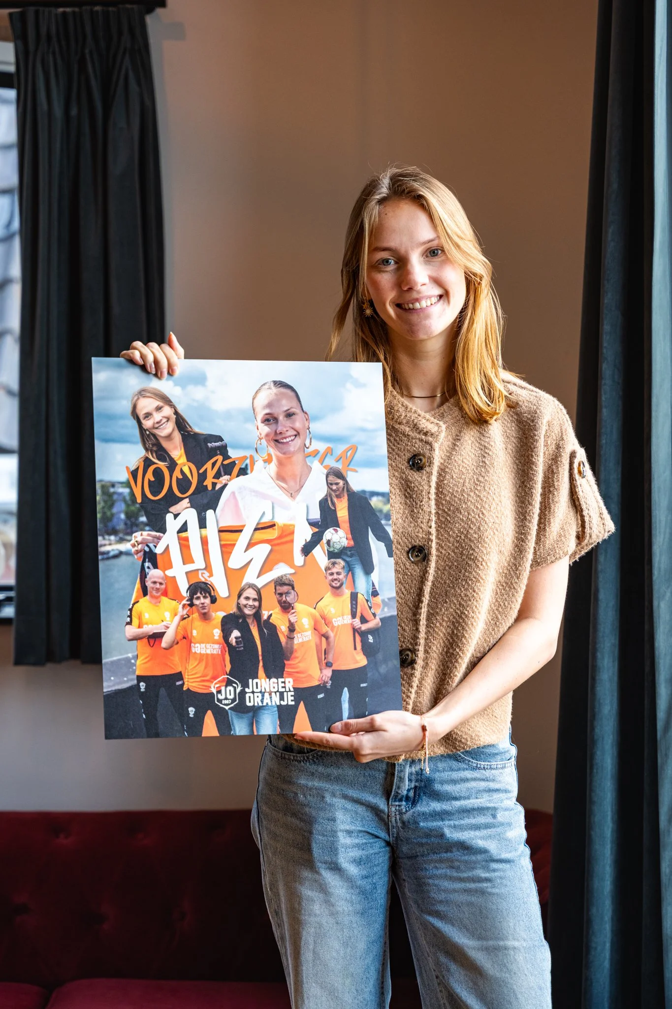 Een jonge vrouw met lang, rood haar glimlacht terwijl ze een poster vasthoudt. Op de poster staan andere mensen in oranje sportkleding, en er is tekst die onder meer 'JONGER ORANJE' bevat.