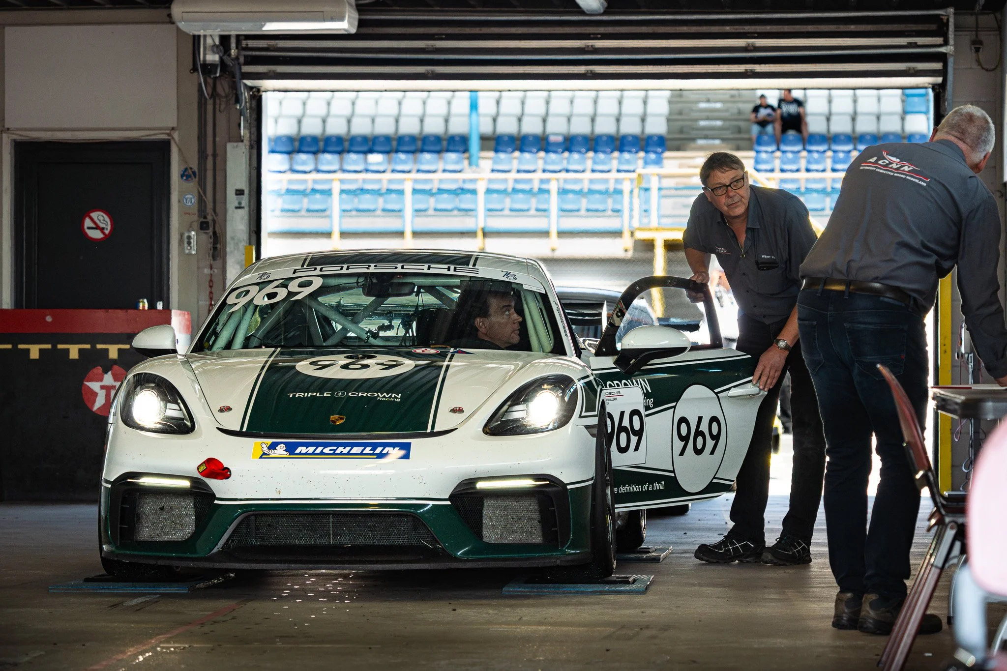 A race car inside a garage with two men preparing it, and an empty grandstand visible in the background.