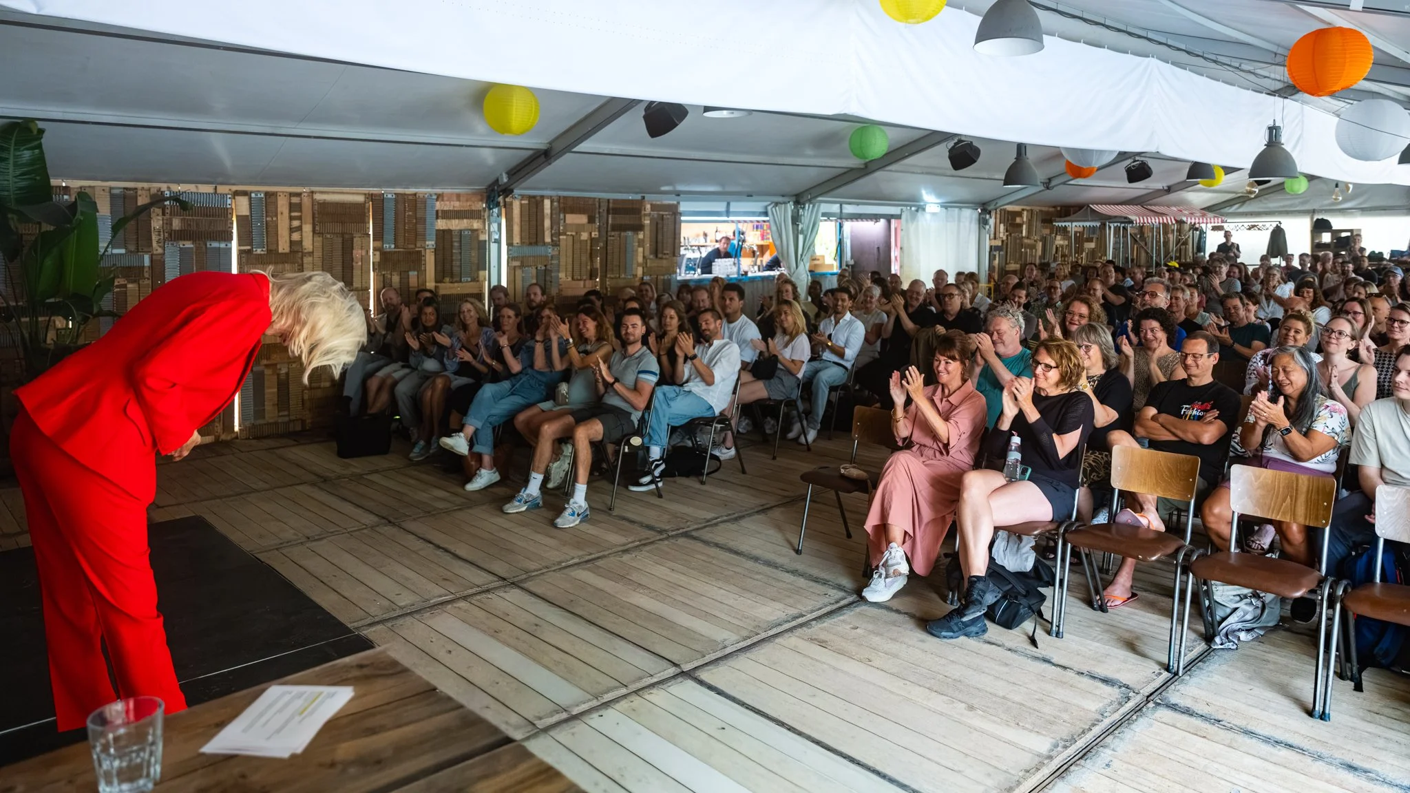 Een vrouw in een rode outfit buigt voorover terwijl ze toeschouwers applaudisseren in een tent met kleurrijke lampionnen en houten decoraties.