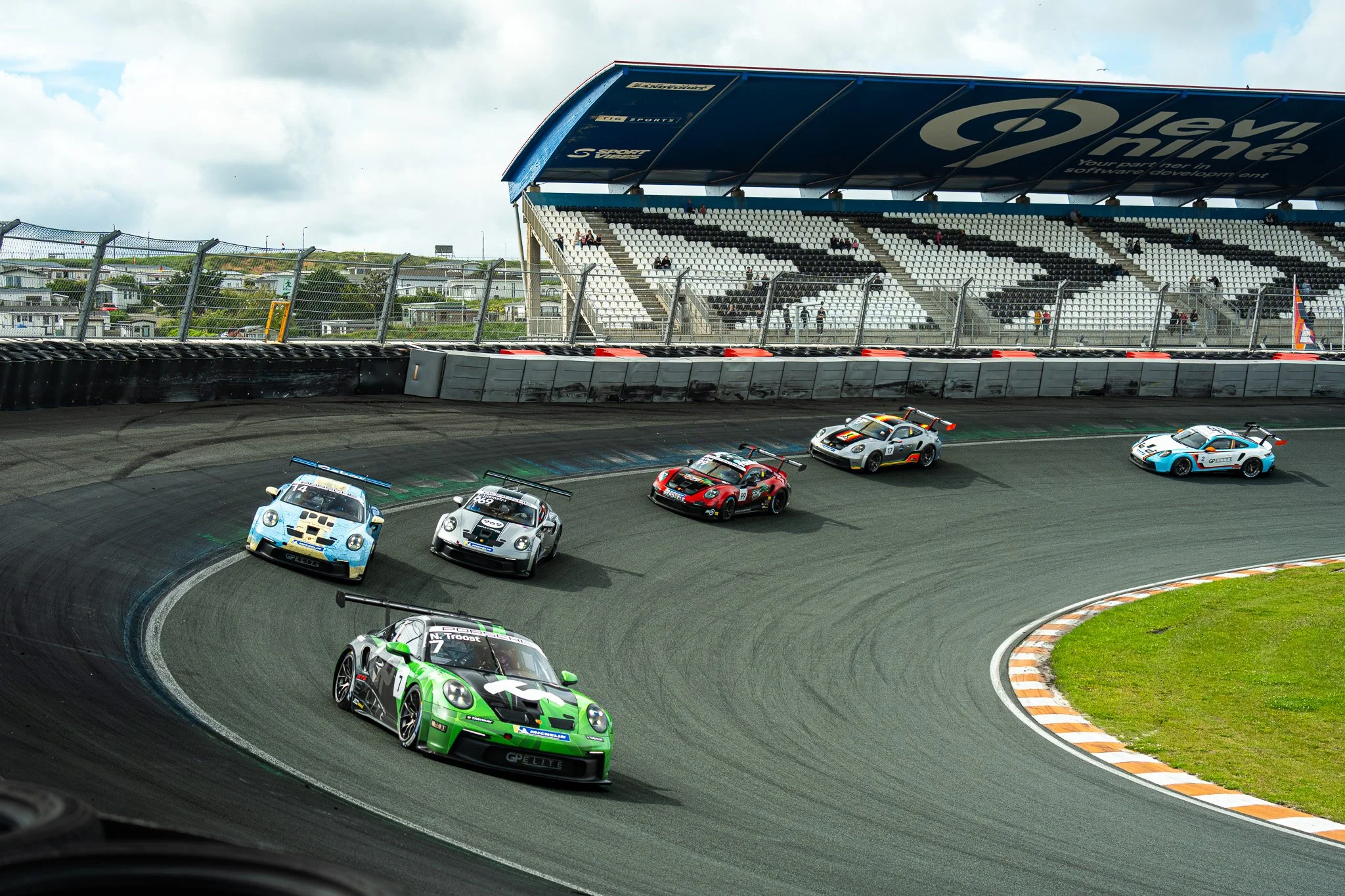 Silhouetten van raceauto's die een bocht nemen op een autosportbaan met een overkapt tribune en een hek aan de rand van de baan.
