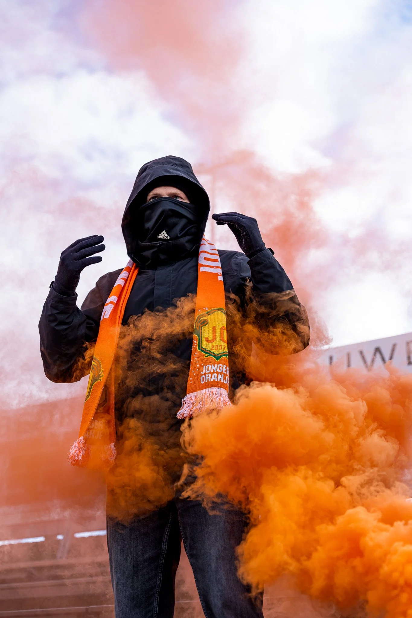 Persoon in zwarte jas, met capuchon en gezichtsschild, omgeven door oranje geurspoor en rook, tijdens demonstratie of protest.