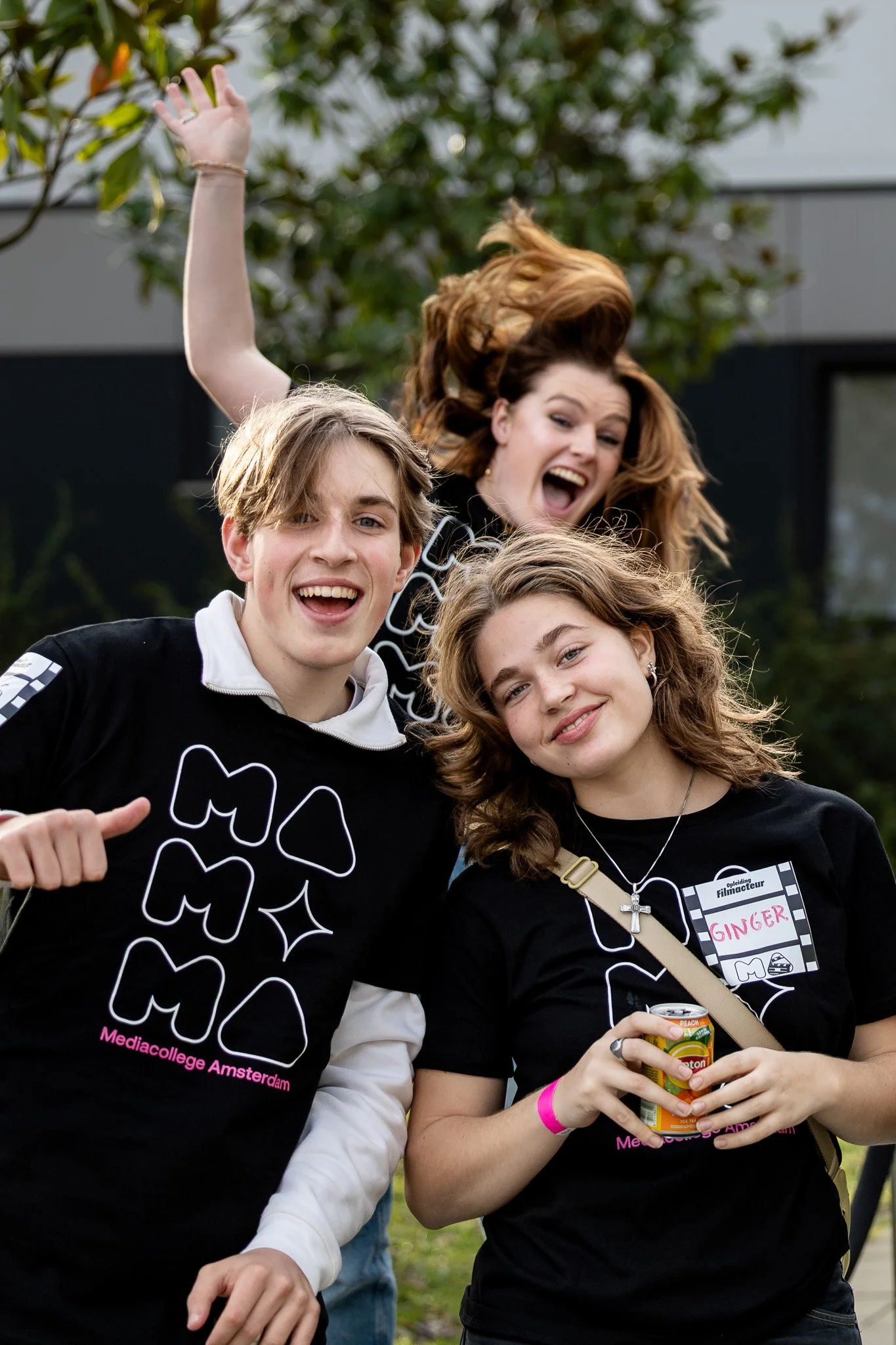 Group of four young people outdoors, smiling, wearing black t-shirts with a white and pink design, one person has a name tag that says 'Ginger', and they appear to be enjoying a social event.