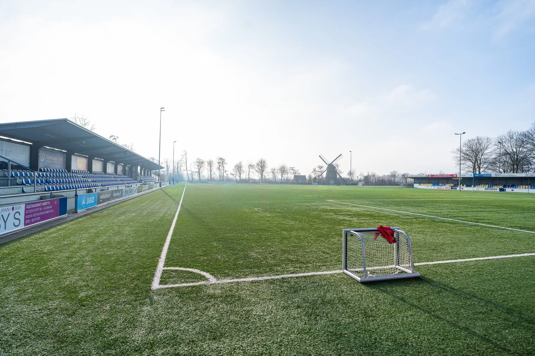 Een leeg voetbalveld met een doel in de voorgrond en tribunes aan de zijkant. In de achtergrond staan windmolens en bomen onder een lichte bewolkte hemel.