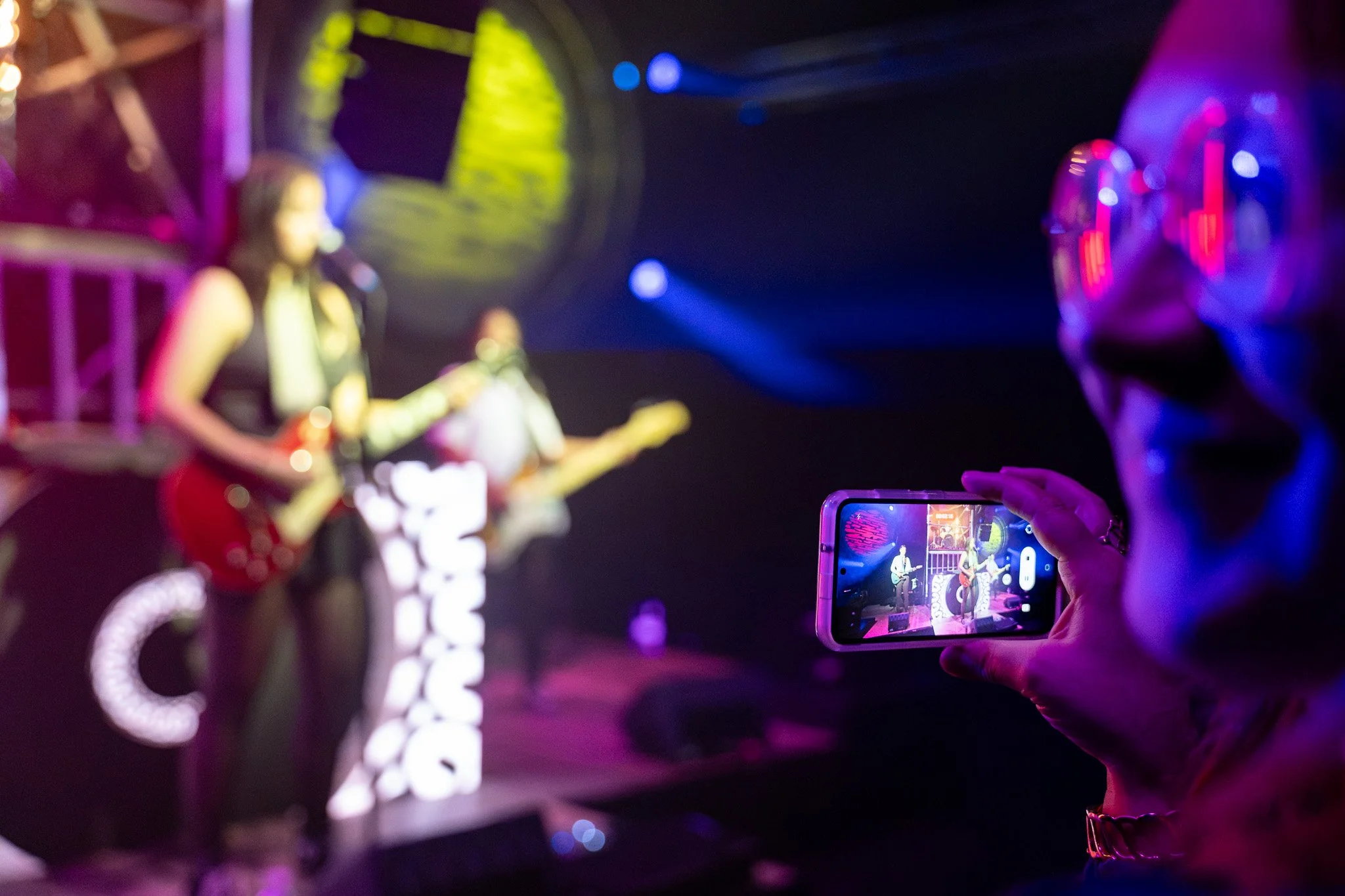 Person taking a photo or video of a live band performing on stage at a concert, with stage lights illuminating the scene.
