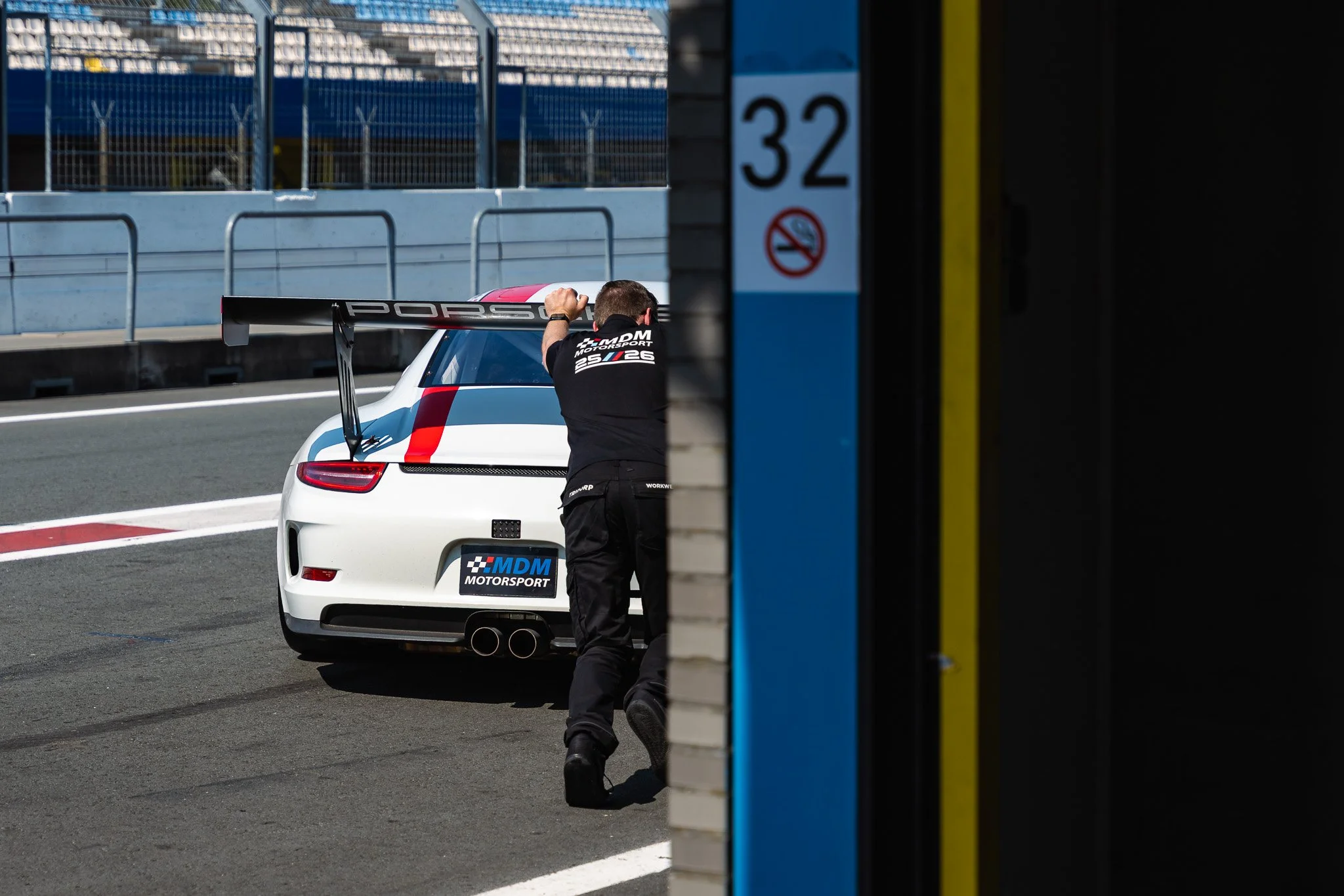 A person in racing gear leaning over a white Porsche race car in the pit lane at a race track, with the pit wall and grandstands in the background.