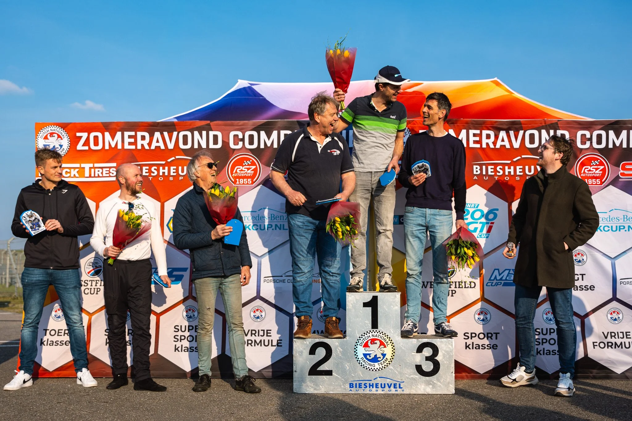 Group of men on a winners' podium celebrating at a racing event, holding flowers and plaques, with a colorful banner and blue sky in the background.
