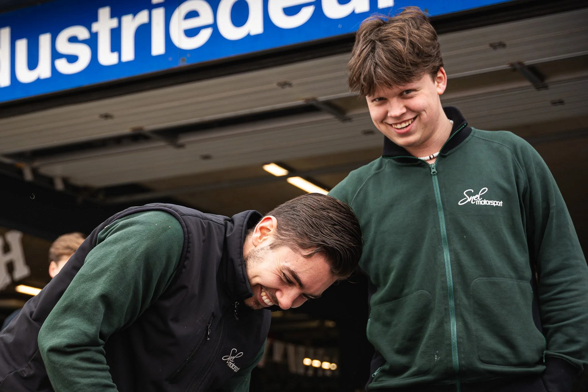 Twee jonge mannen lachen en lachen samen onder een overkapping met een bord met de tekst 'Industriec...' in de achtergrond.