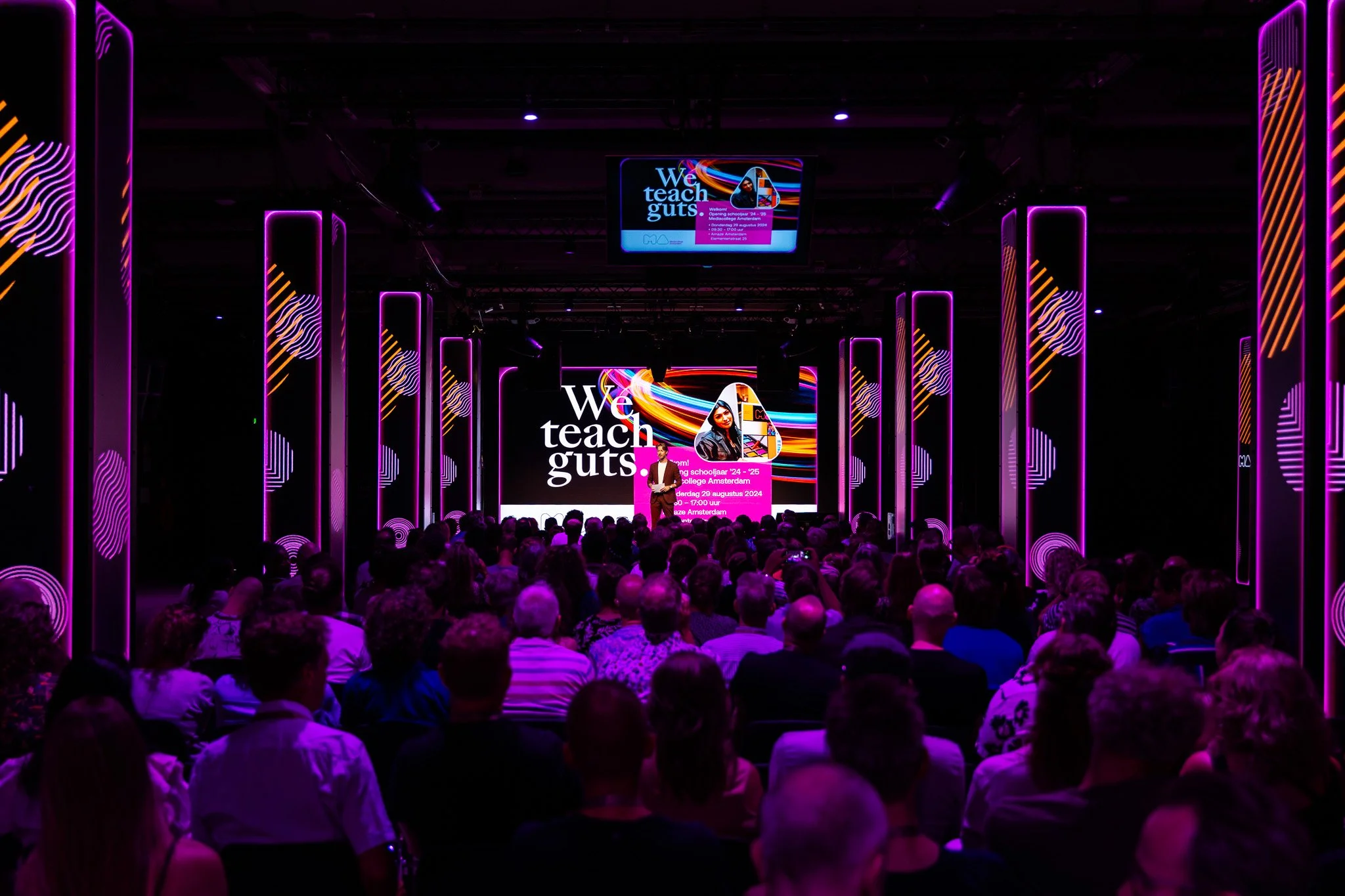 A speaker presenting at a conference called 'We teach guts' in a dark room with purple neon lights, a large screen behind them displaying event details and a photo of a woman, and an audience seated and listening.