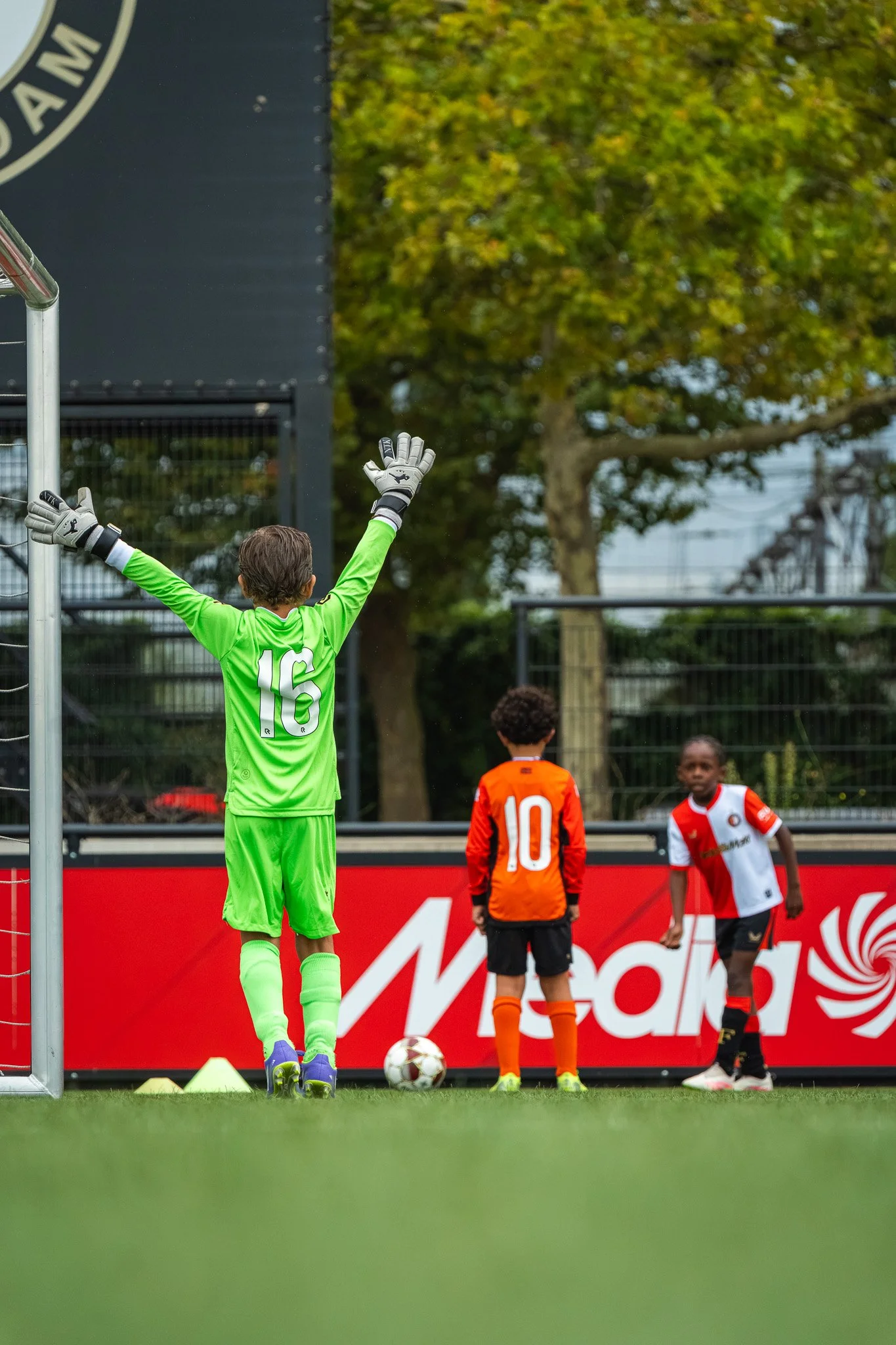 Kinderen op een voetbalveld, met een goalkeeper in een groen tenue met nummer 16, klaar om een penalty te nemen. Anderen staan klaar achter de lijn, met een bal en gele konen op de grond.