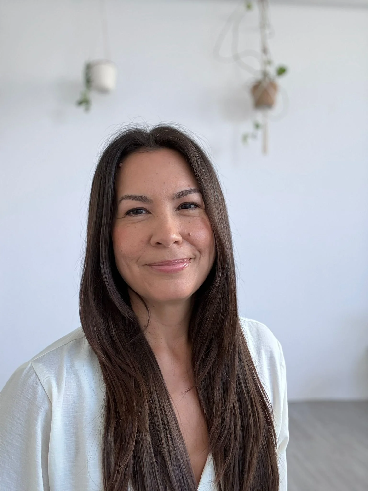 A woman with long brown hair and light skin smiling in front of a plain white wall with hanging plants visible at the top.
